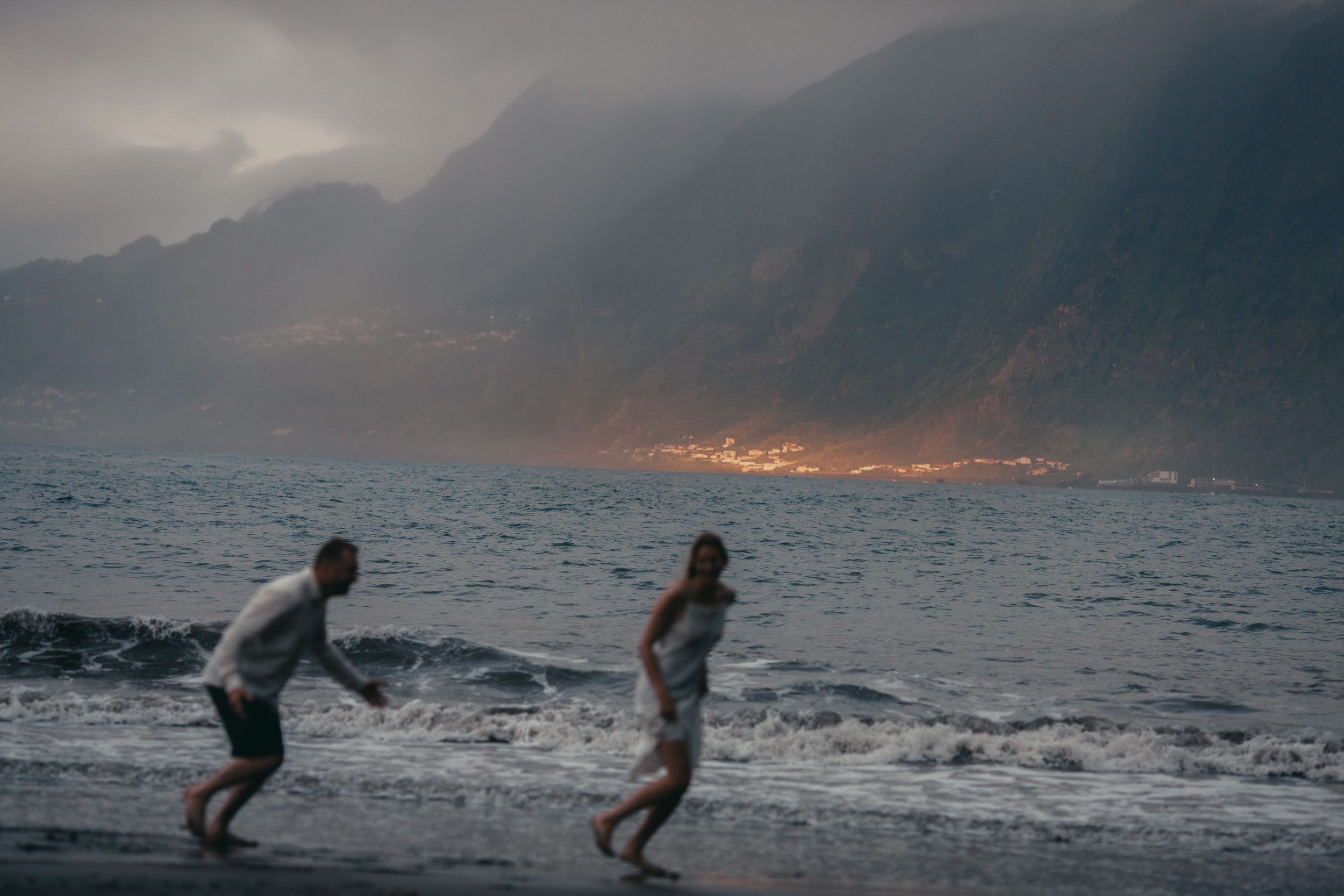 Sunset silhouette of a couple on a dark beach, fog drifting over the green ridges, north Madeira