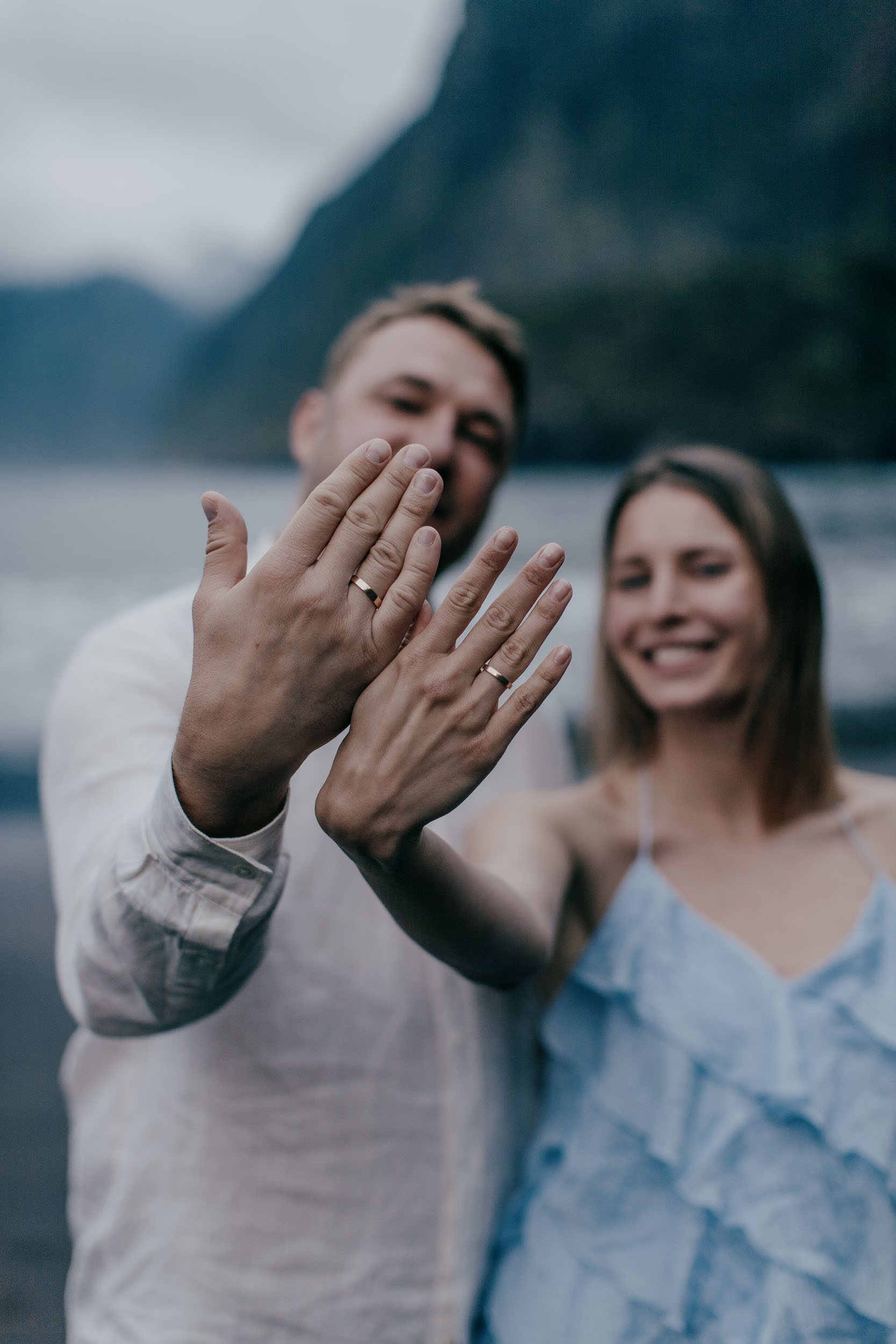 Two hands held up close together with wedding rings on each, faces softly out of focus behind, dramatic green cliffs, Madeira