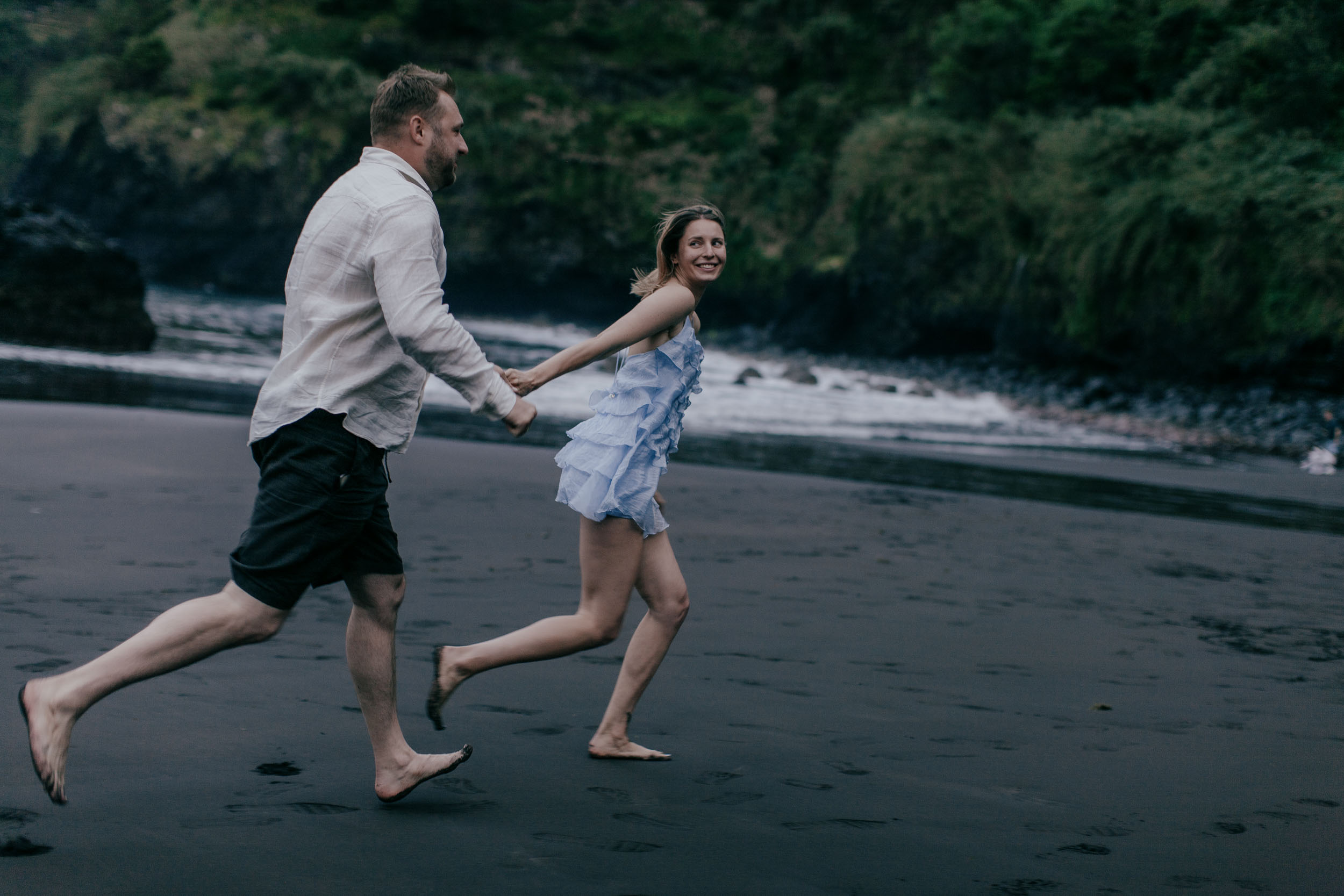 Couple running together on a black sand beach hand in hand, she looks back smiling in a pale blue dress, north Madeira coast