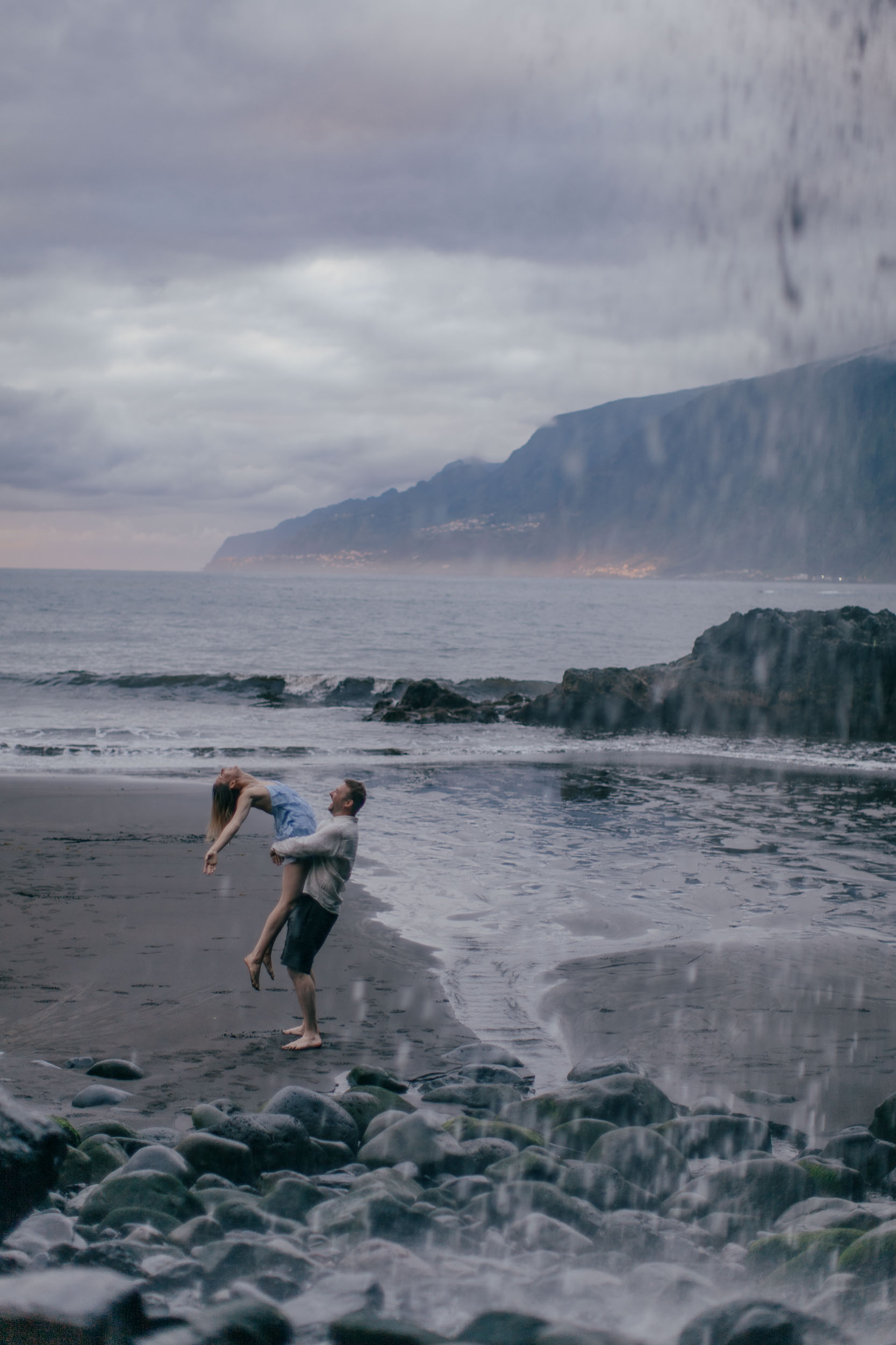 Couple on a black sand beach, she in a pale blue dress with ruffled bodice, he in white linen, Madeira
