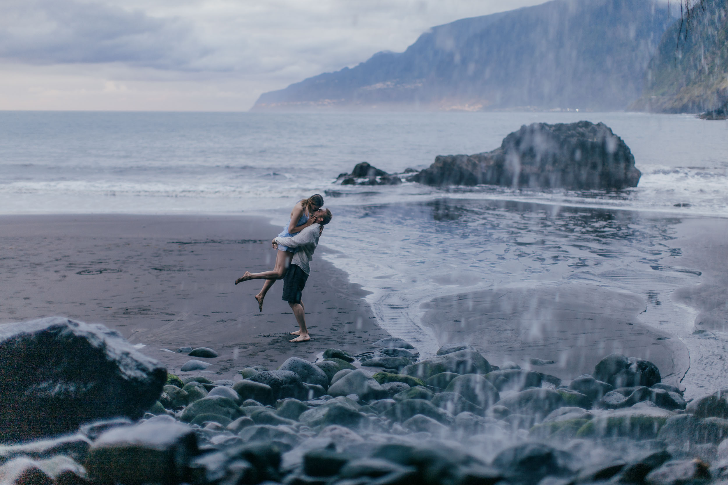 Couple holding hands on dark sand, pale blue dress catching the wind, north Madeira coast
