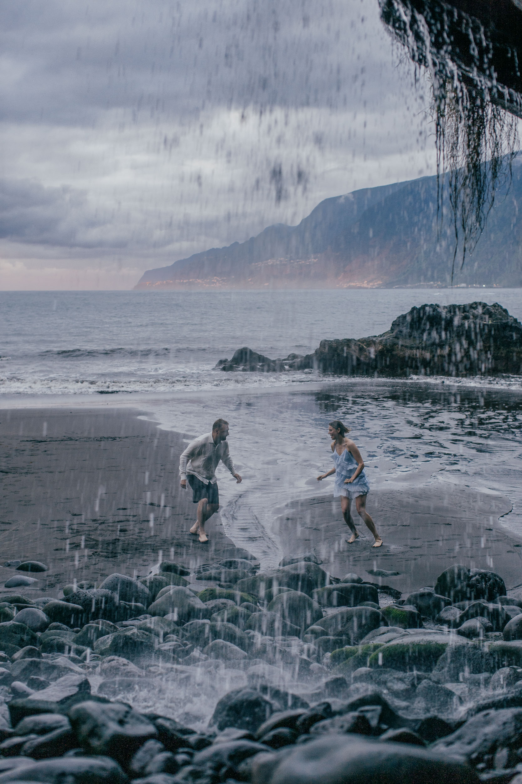 She in blue, he in white, walking on a black sand beach at the foot of dramatic cliffs, Madeira