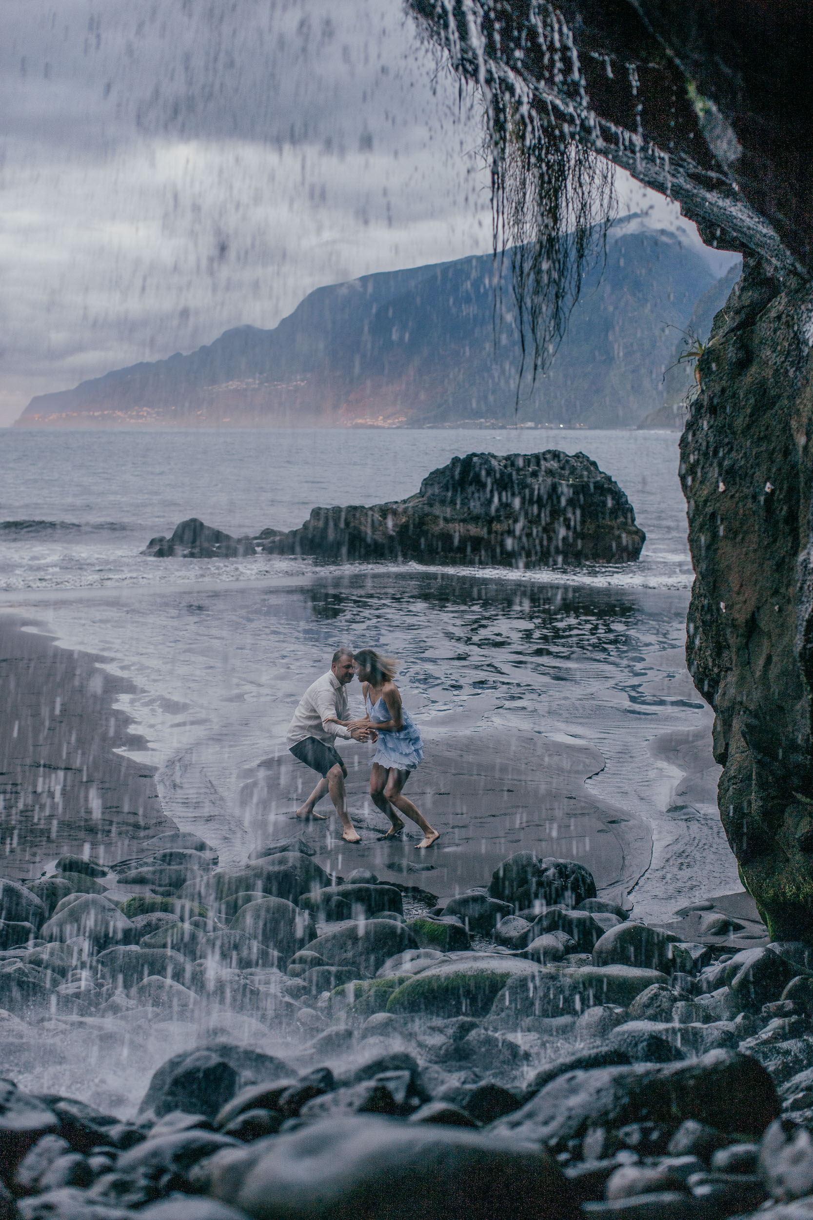 Couple running together on a black sand beach, she laughing back at the camera in a blue dress, Madeira