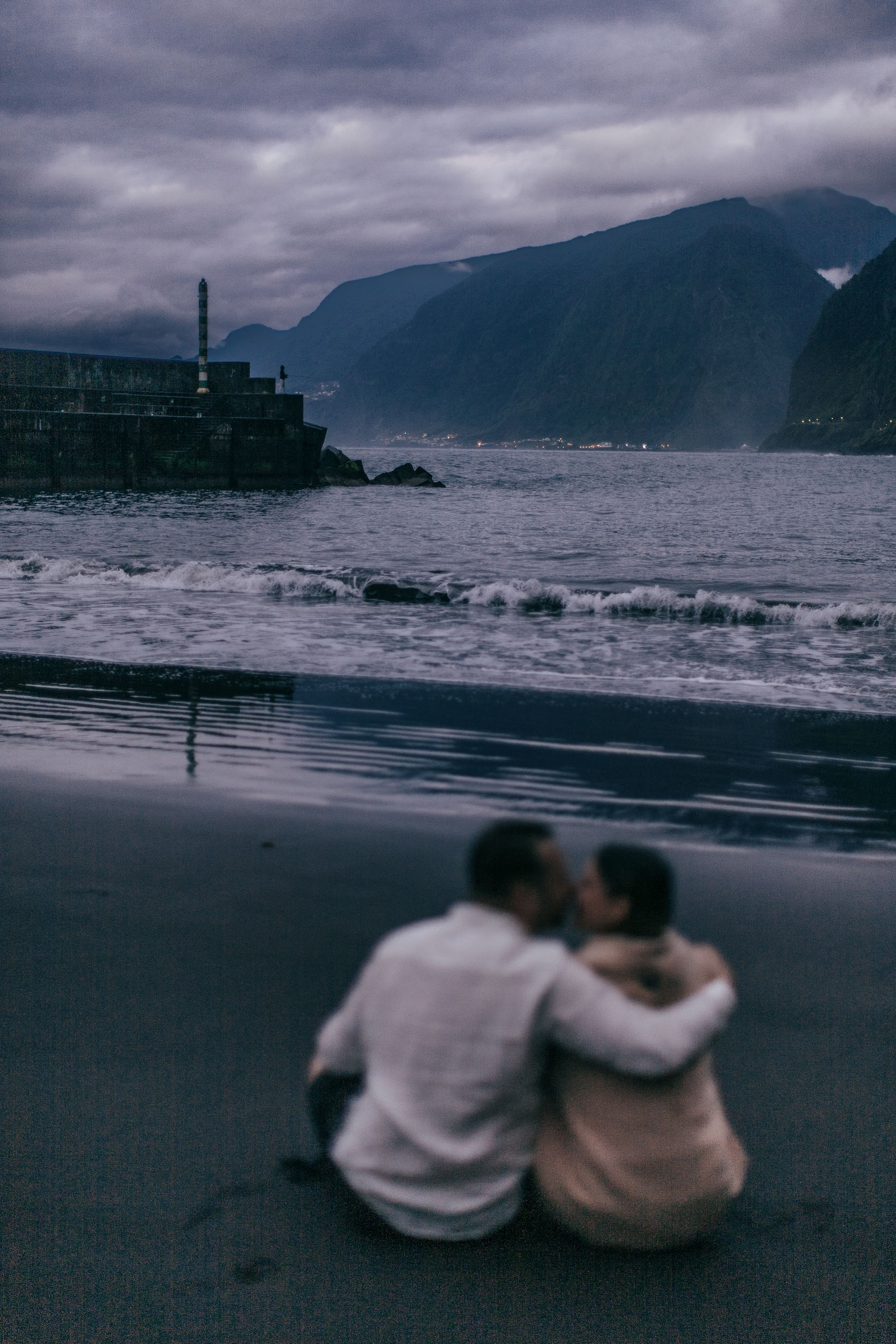 Couple at last light on a black sand beach, dramatic cliffs filling the background, Madeira