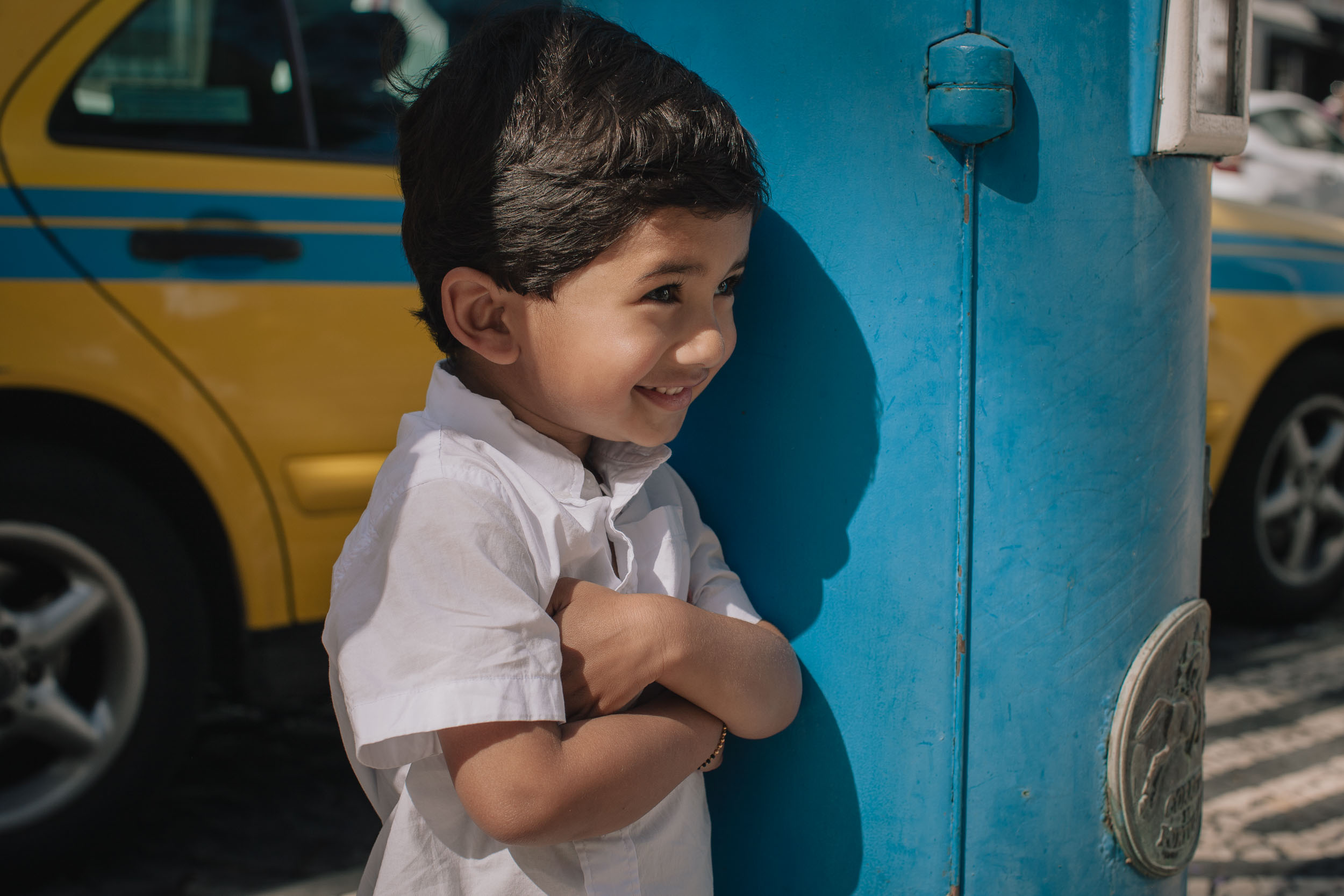 Young boy smiling with arms crossed, leaning against a blue painted door on a Funchal street, yellow taxi visible behind, Madeira