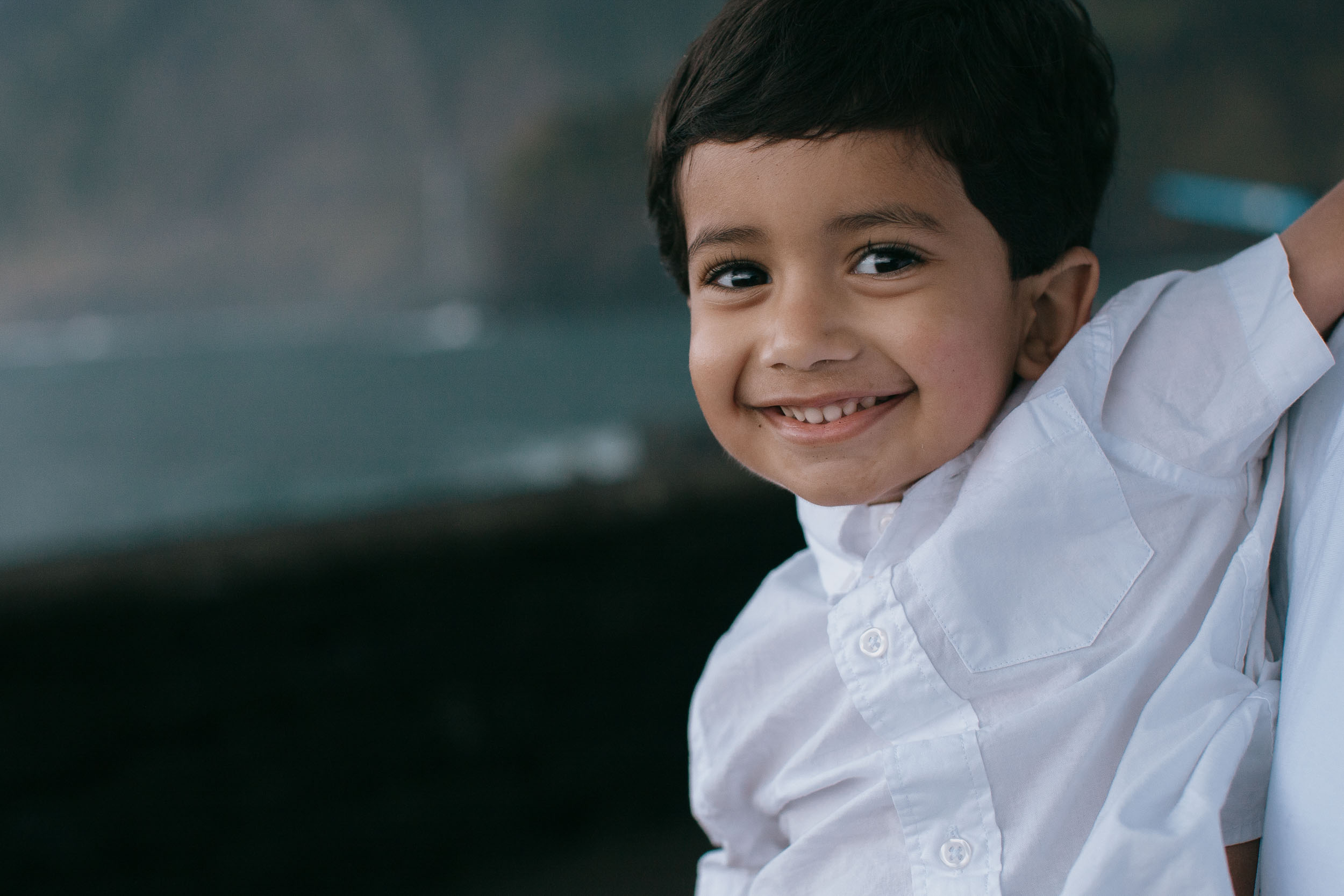 Close-up of boy grinning broadly at camera, white shirt, dark volcanic rocks and moody sea behind, Madeira