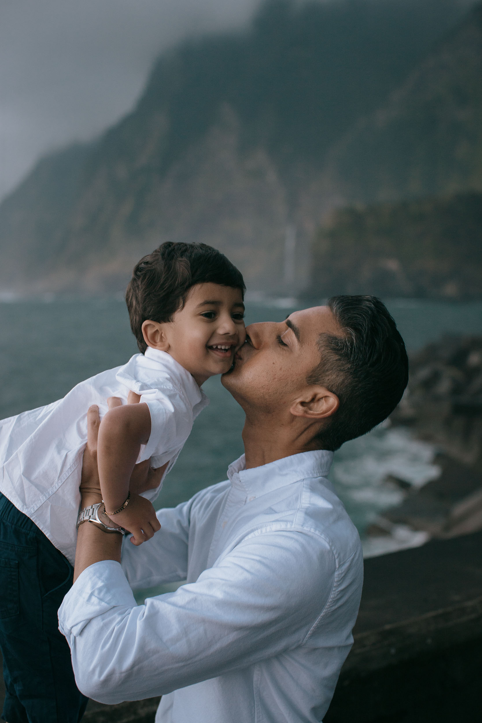 Father lifting and kissing laughing son's cheek, dramatic green cliffs with waterfall behind, moody overcast sky, north Madeira