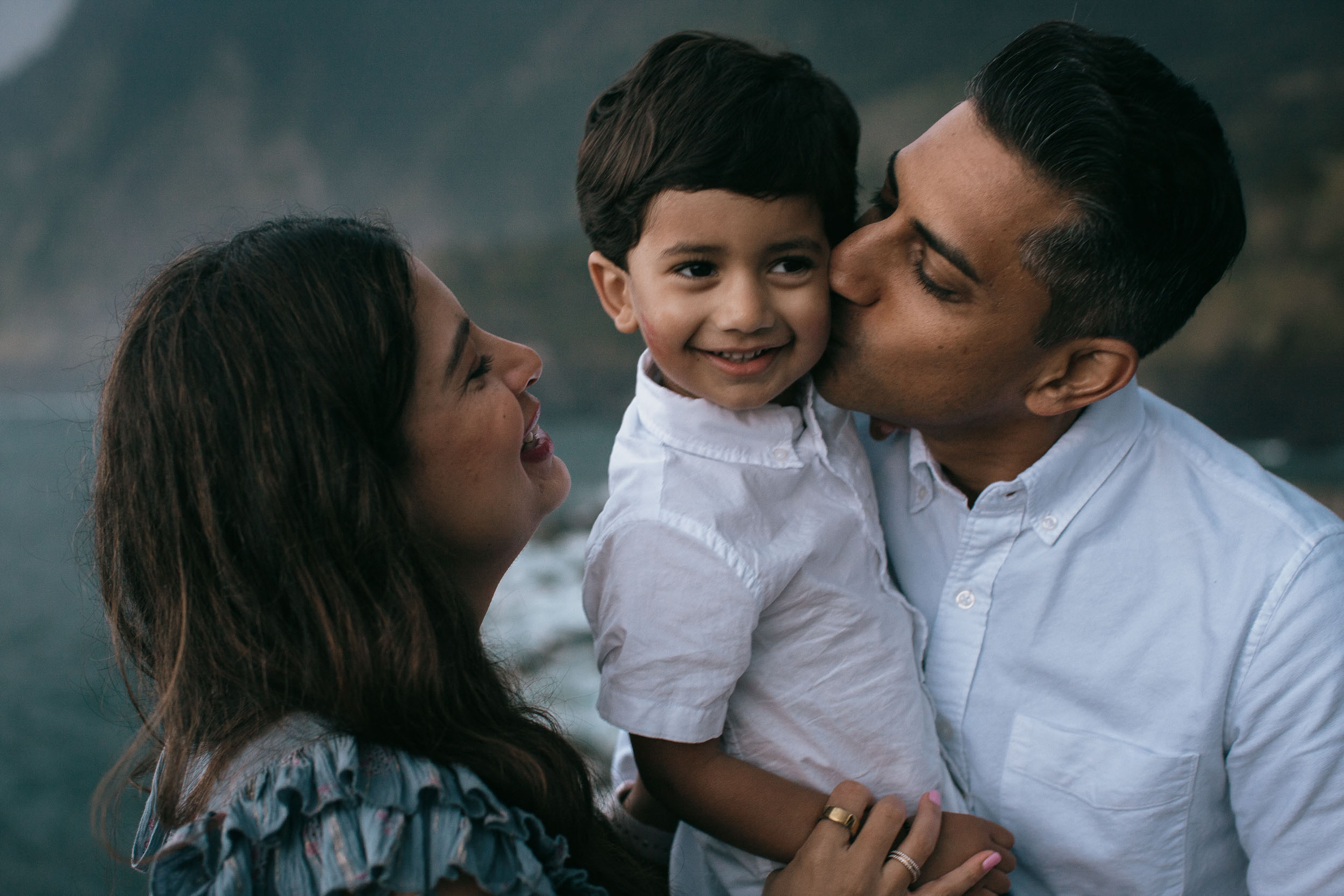 Both parents kissing boy's cheeks simultaneously, boy smiling at camera, moody dramatic cliffs behind, north Madeira coast