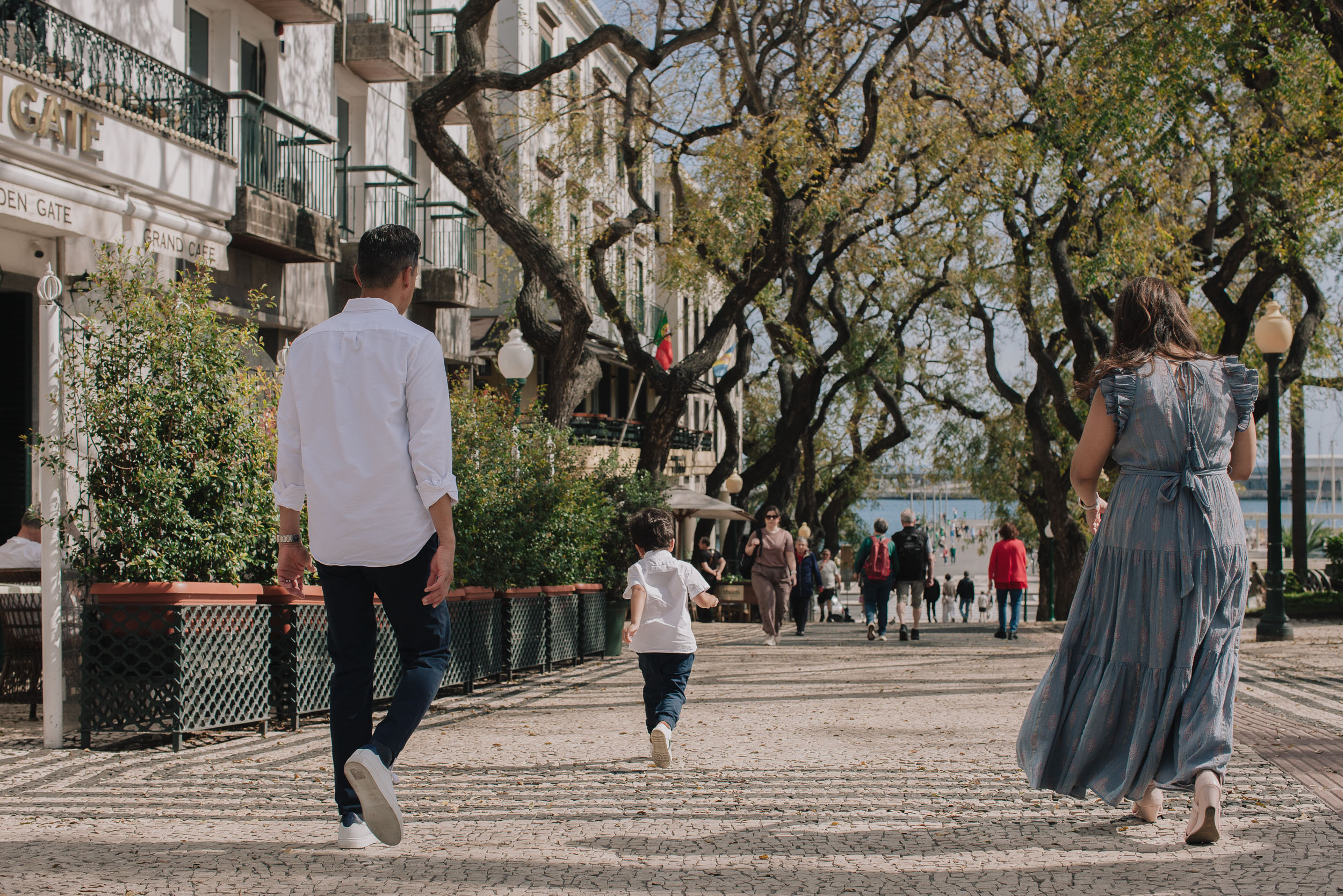 Family walking along tree-lined Funchal promenade, backs to camera, boy running ahead, Golden Gate hotel visible, Funchal marina glimpse, Madeira