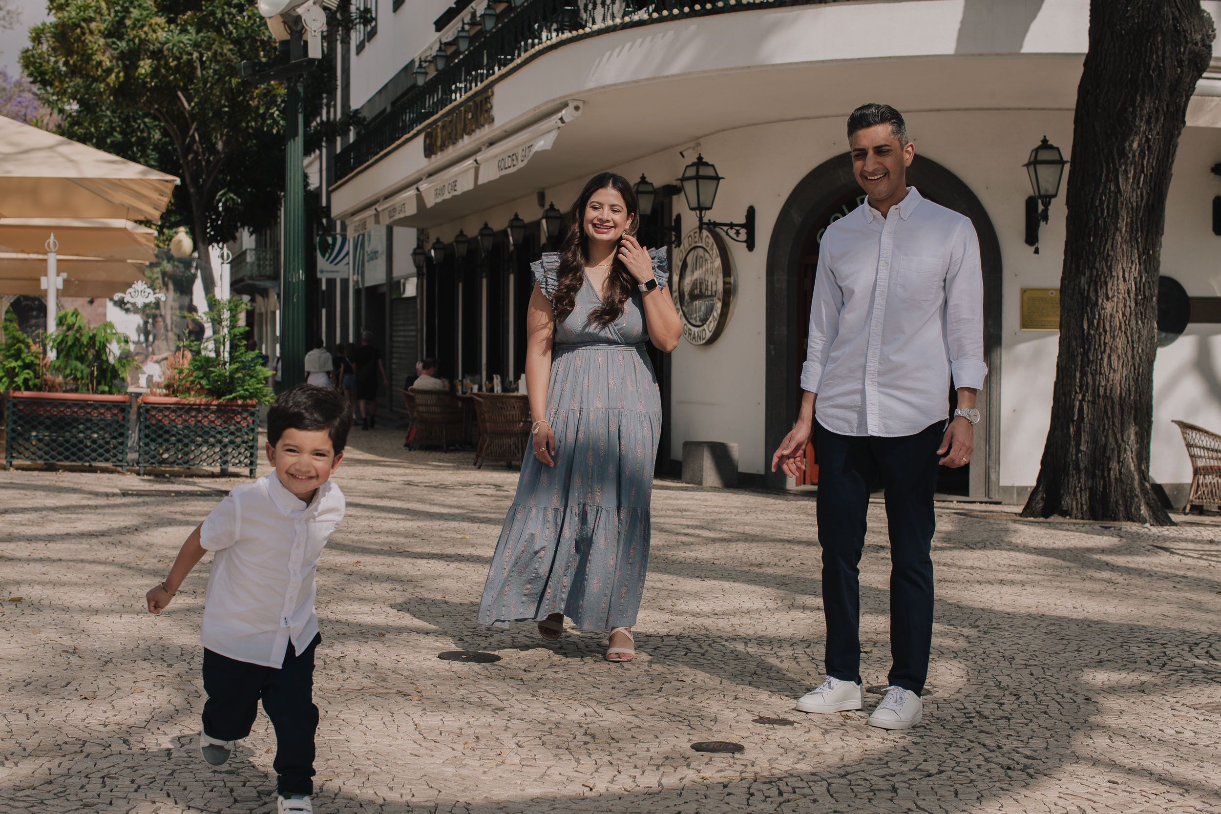 Boy running ahead laughing on Funchal cobblestone plaza, parents following and smiling, colonial building with arched facade behind, Madeira