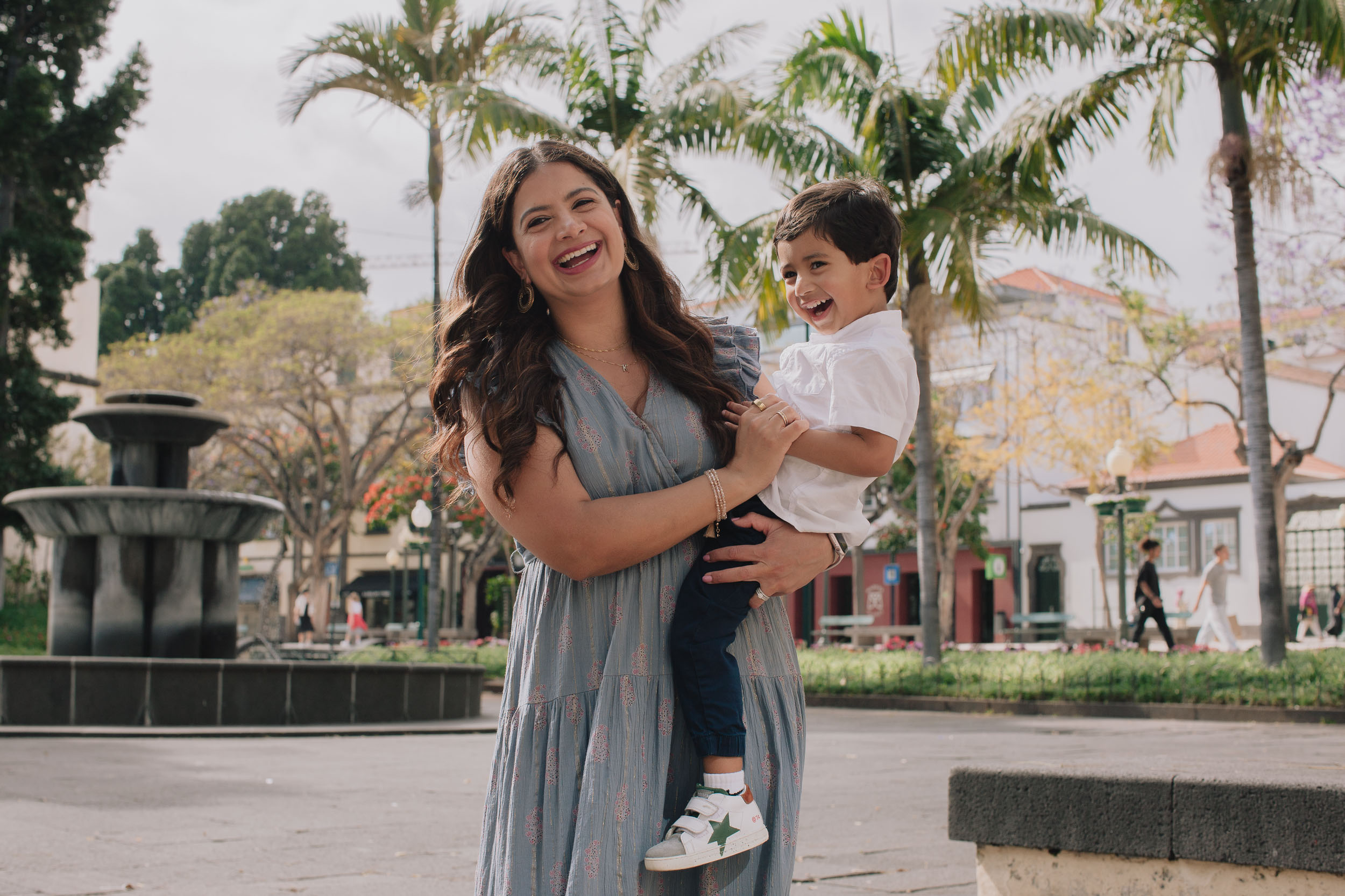 Mother laughing as she lifts boy up, he laughing too, Funchal park garden with stone fountain and palm trees behind, Madeira