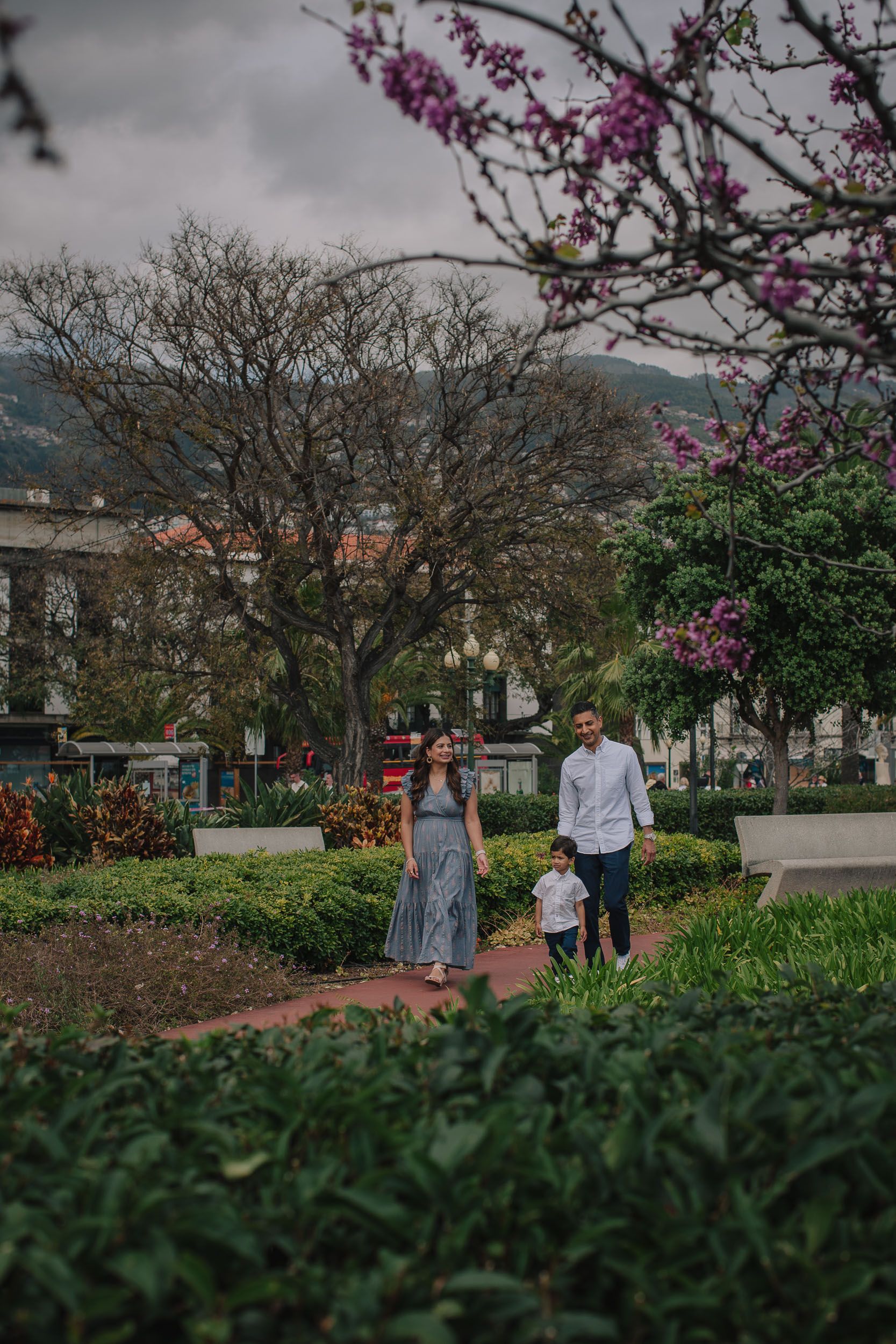 Boy in white shirt flexing arms and grinning at camera on Funchal cobblestone promenade, city behind, mother visible in background, Madeira