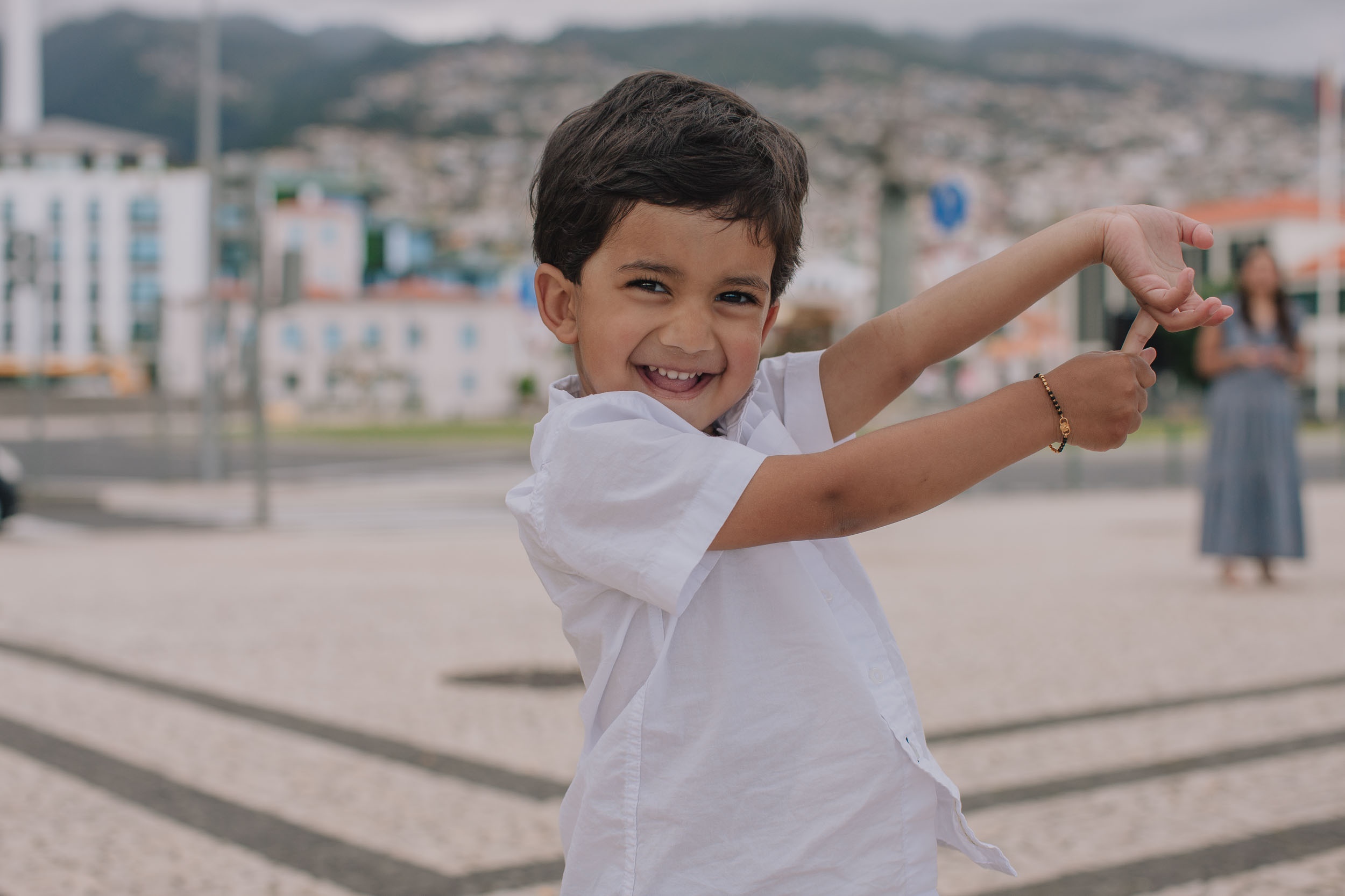 Boy dancing and posing on Funchal waterfront cobblestone promenade, huge grin at camera, mother in background, Funchal city behind, Madeira