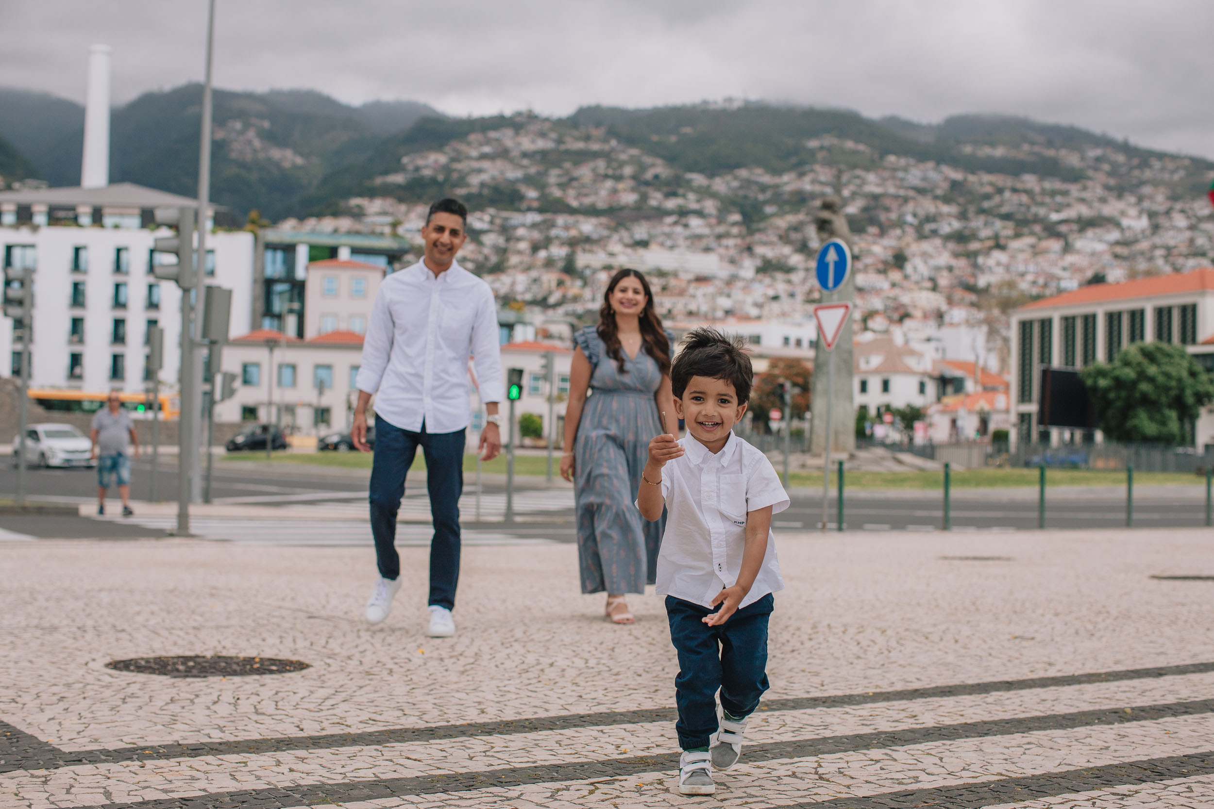 Family of three walking on Funchal cobblestone square, boy running toward camera laughing, parents behind, Funchal hill and city behind, Madeira