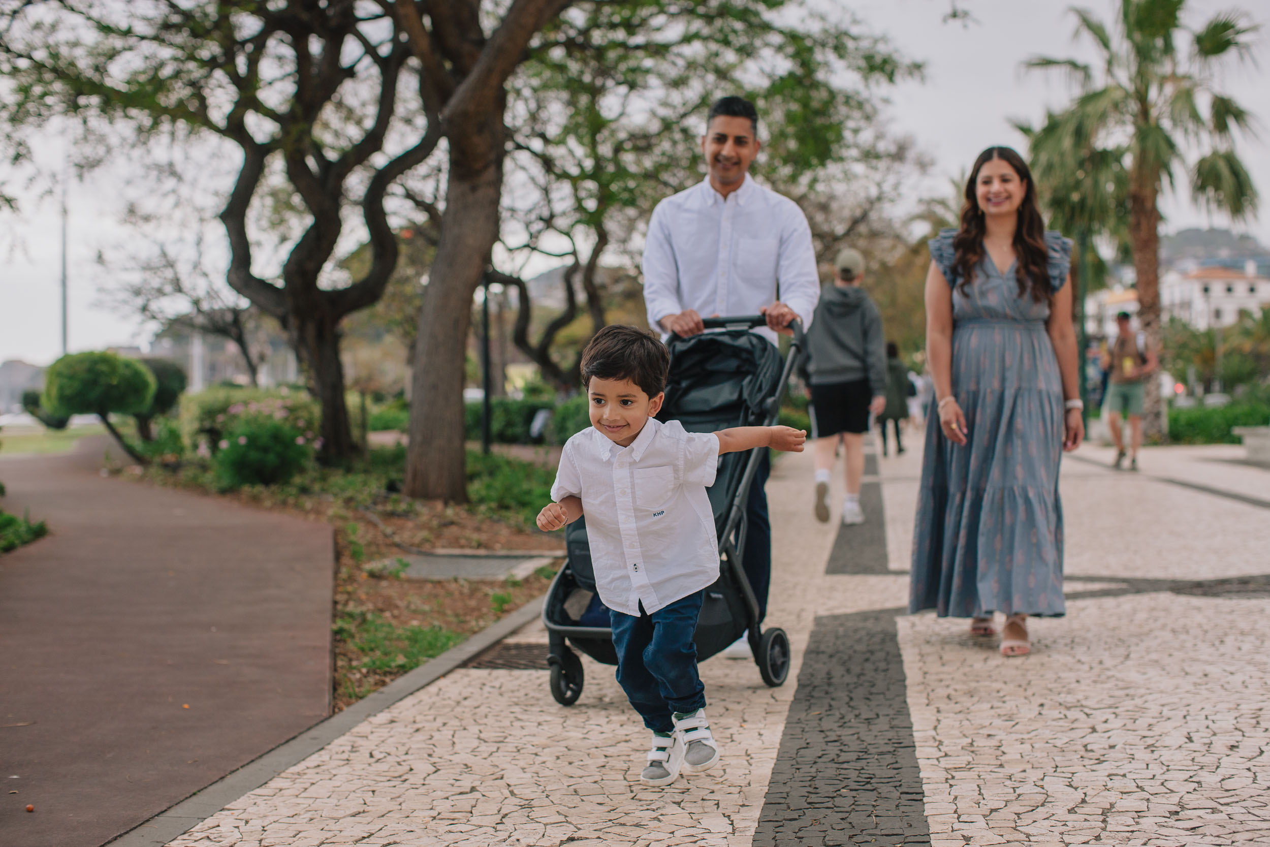 Father pushing stroller while boy runs ahead with arms out, mother smiling behind, Funchal tree-lined promenade path, Madeira