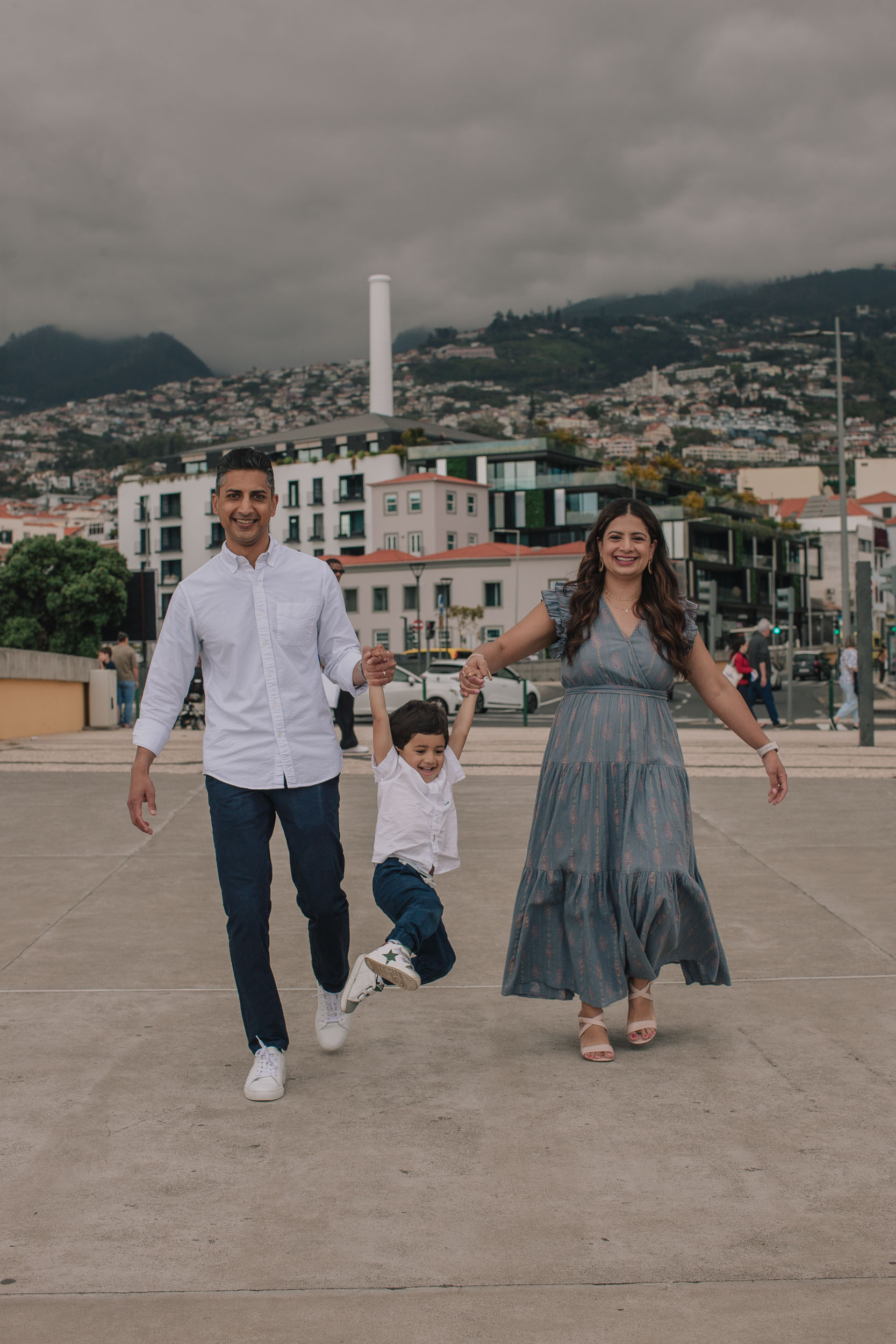 Family of three holding hands swinging boy between them on Funchal waterfront plaza, Funchal city and chimney behind, overcast sky, Madeira