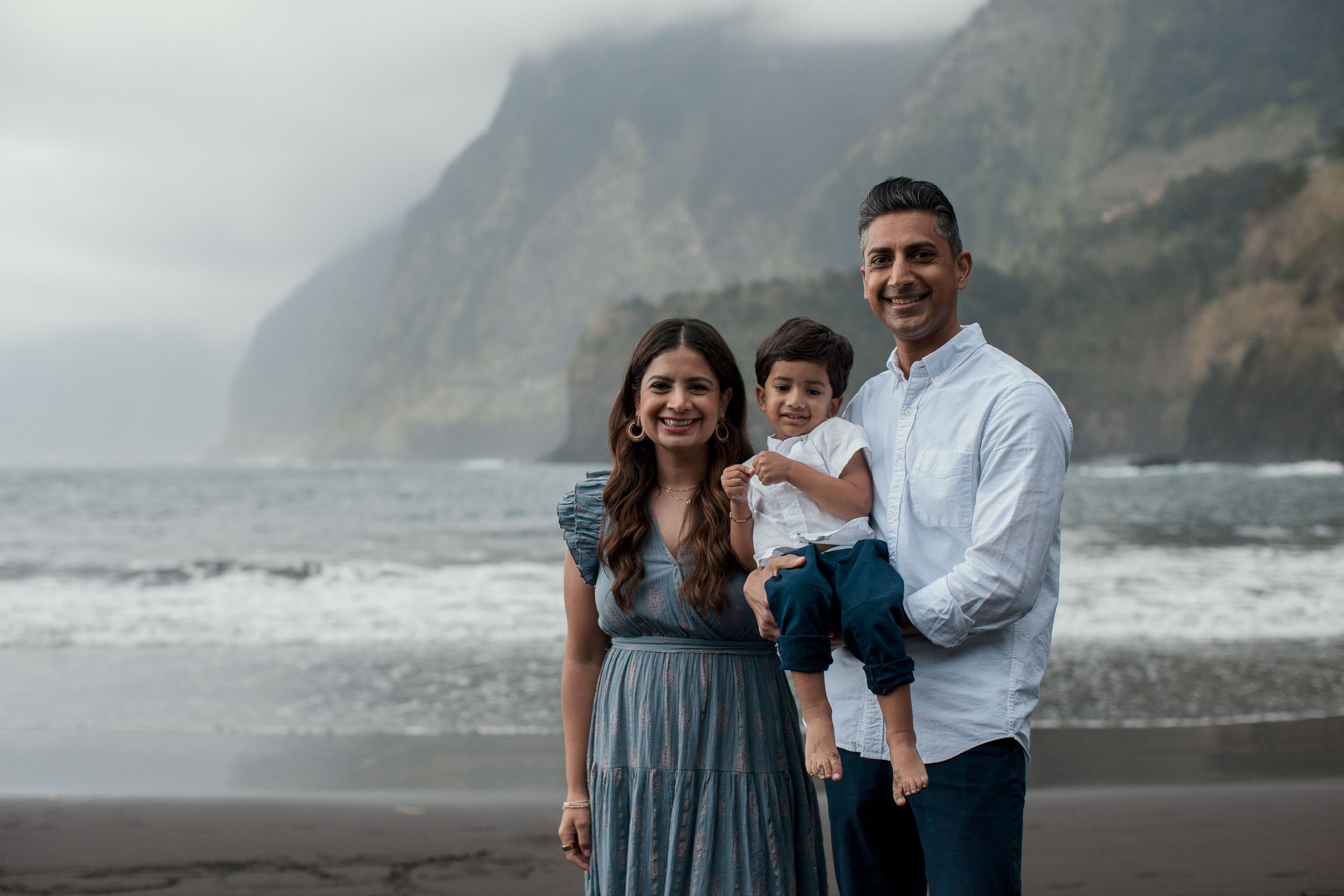 Family portrait at Seixal. mother in blue dress, father holding son, dramatic misty Seixal cliffs and black sand beach behind, Madeira