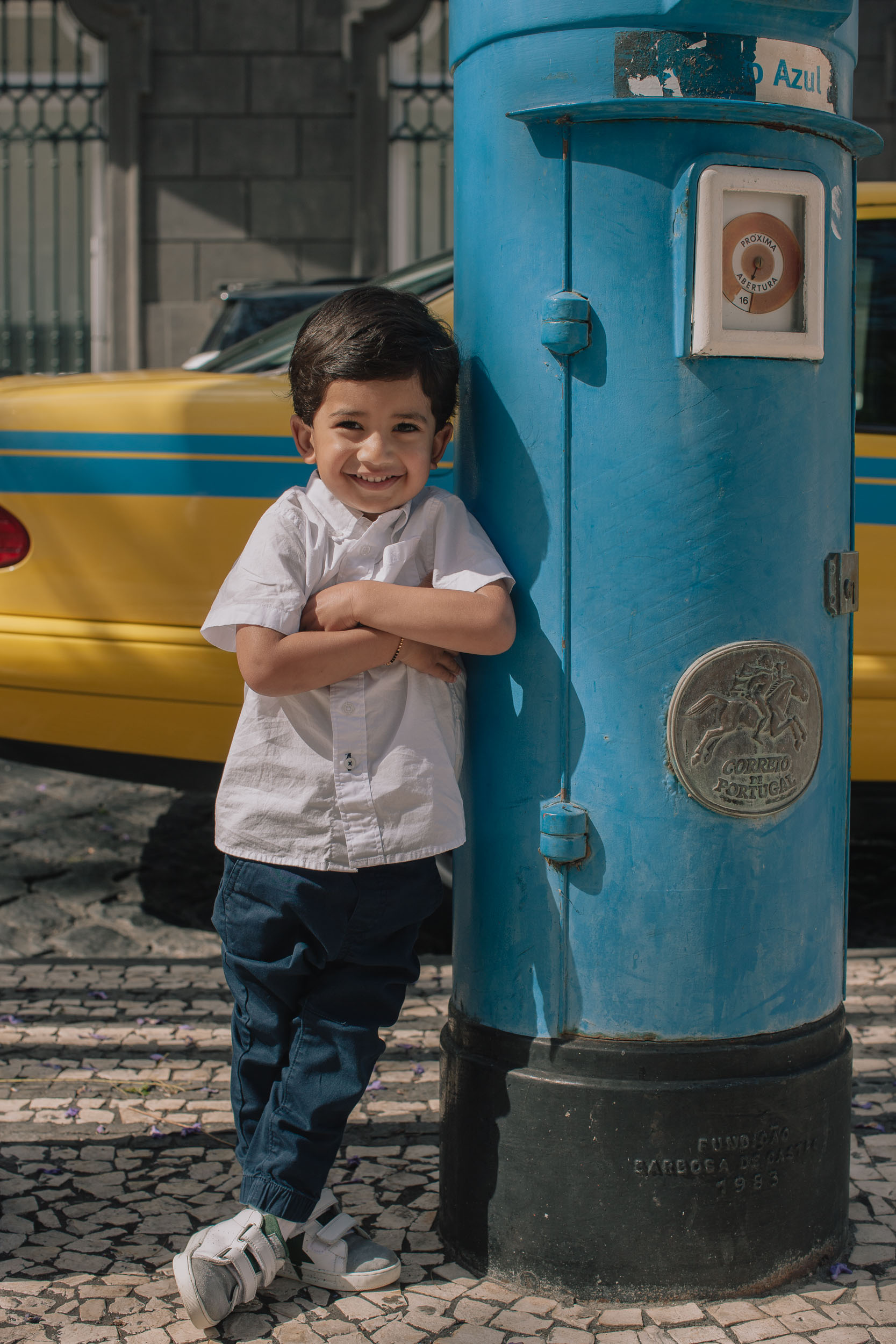 Boy full body leaning against a blue Portuguese letter box with arms crossed smiling, yellow Funchal taxi behind, cobblestone street, Madeira