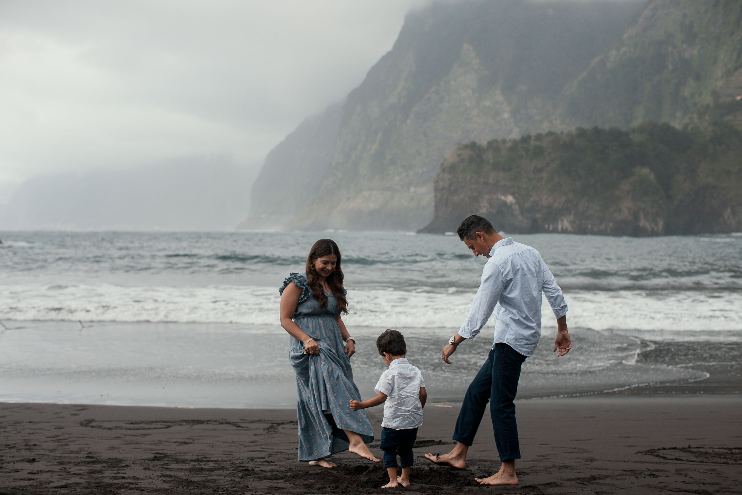 Family of three on Seixal black sand beach, parents watching over boy at the surf line, dramatic misty cliffs behind, Madeira