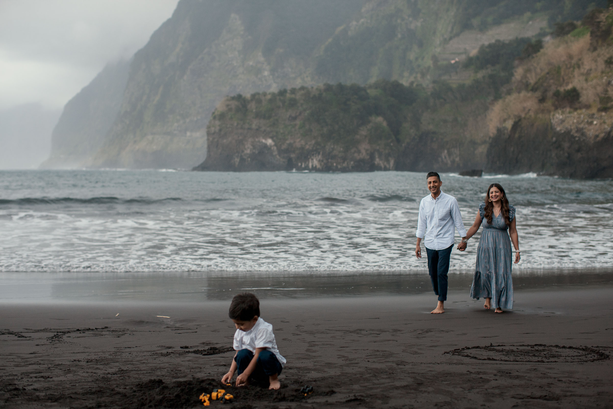 Boy playing with toy cars in black Seixal sand in foreground, parents walking hand in hand behind, dramatic misty cliffs and Atlantic, Madeira