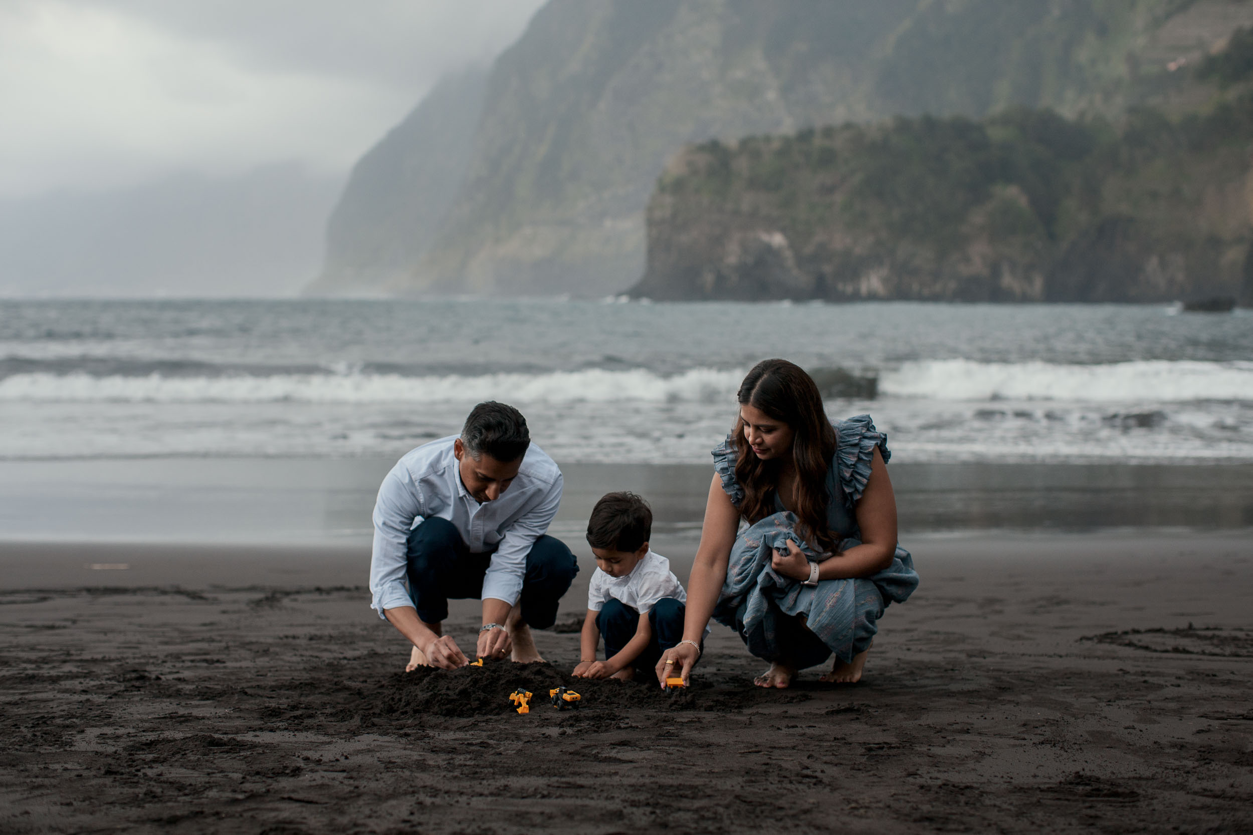 Family of three holding hands on Seixal black sand beach, facing the Atlantic ocean and cliffs, Madeira