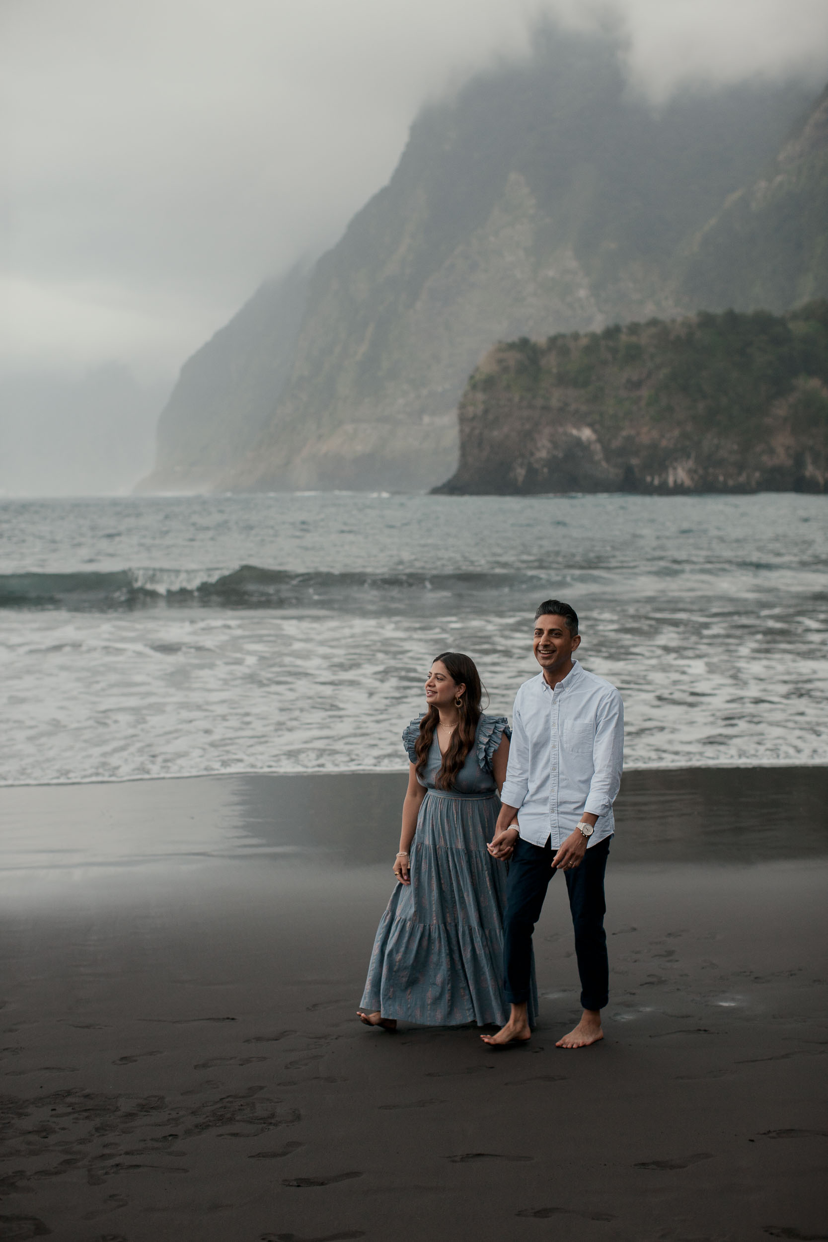 Parents walking hand in hand barefoot on Seixal black sand beach, misty dramatic cliffs behind, Atlantic waves, Madeira