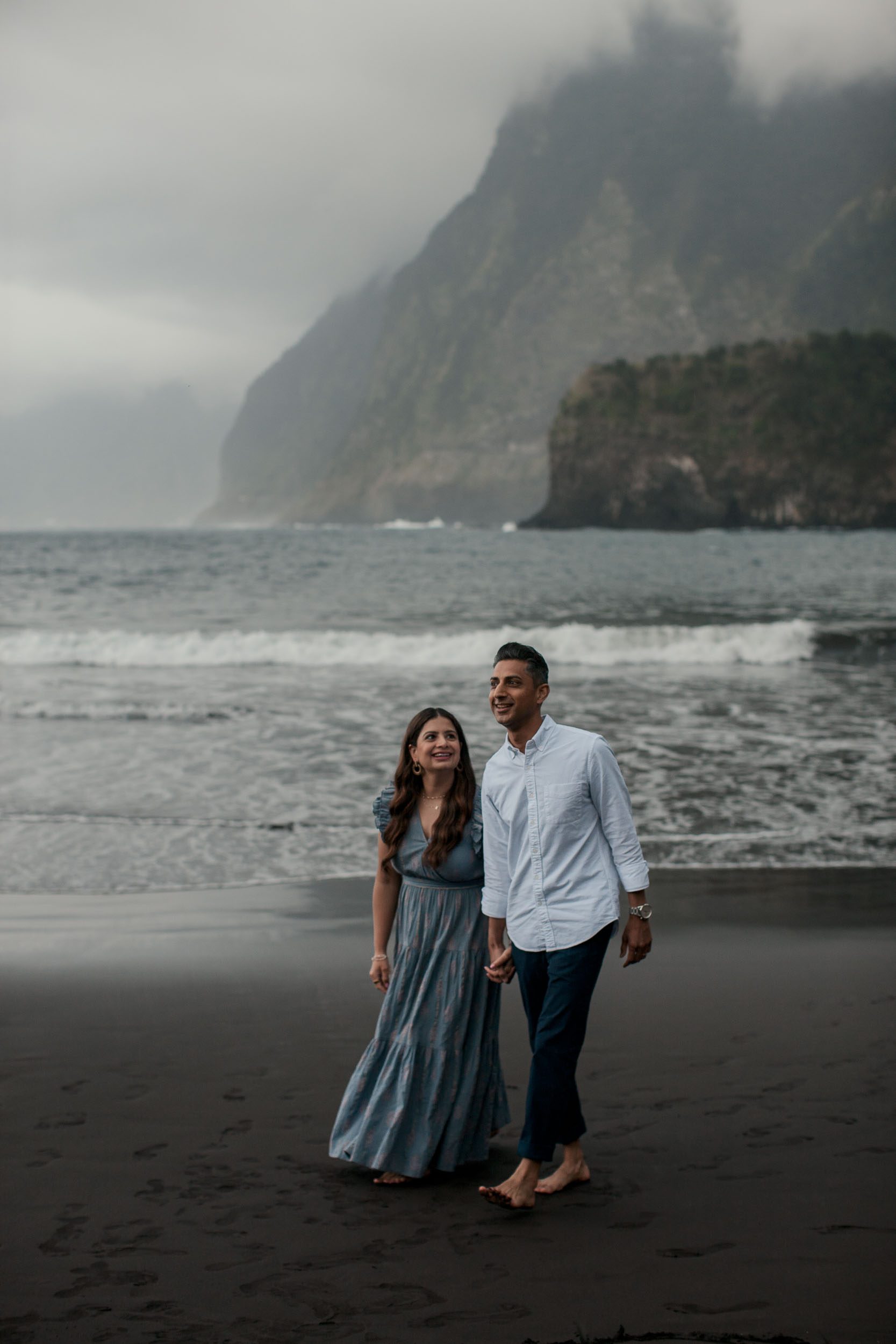 Parents walking hand in hand on Seixal black sand beach, woman looking up laughing, misty Seixal cliffs and Atlantic behind, Madeira