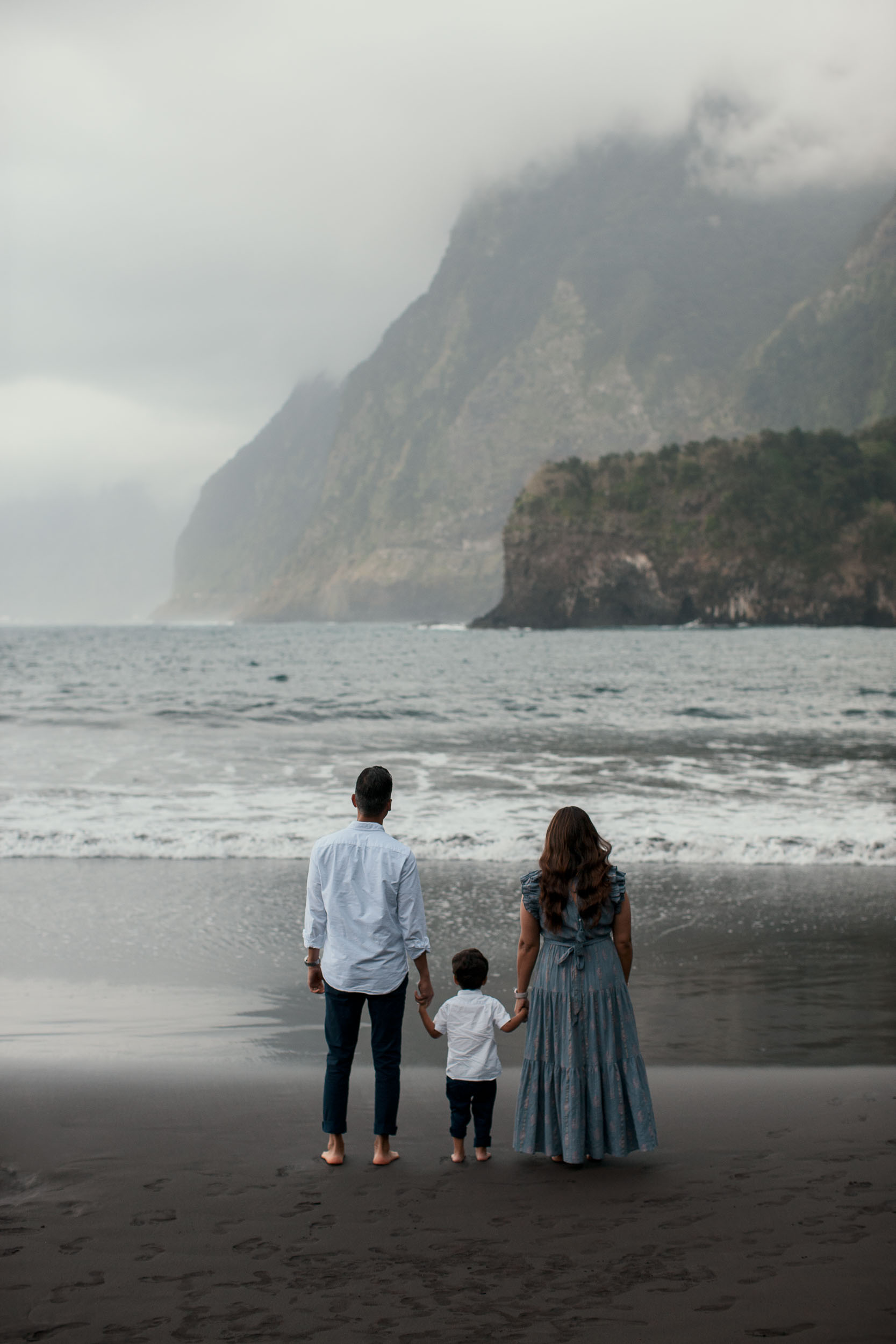 Family of three from behind holding hands, facing the Atlantic and dramatic misty Seixal cliffs, black sand beach, Madeira