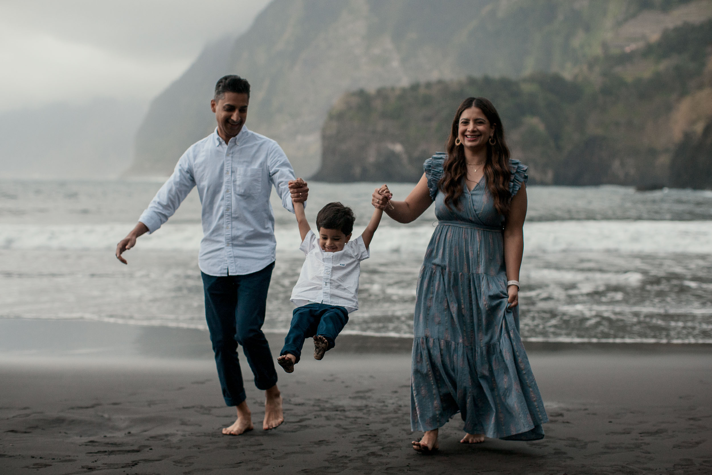 Parents swinging laughing boy between them on Seixal black sand beach, dramatic cliffs and surf behind, Madeira