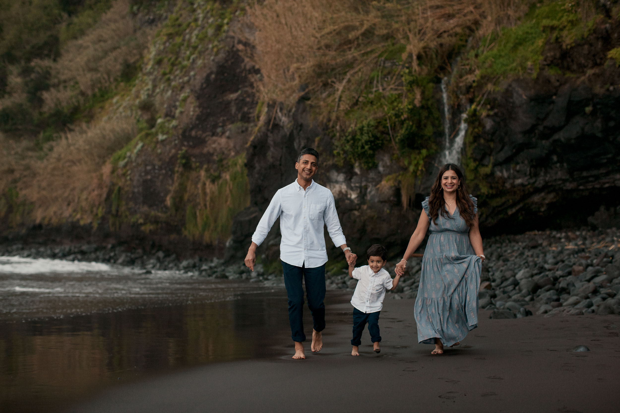 Family of three walking along Seixal black sand shoreline, small waterfall on the cliffside behind, boy between parents, Madeira