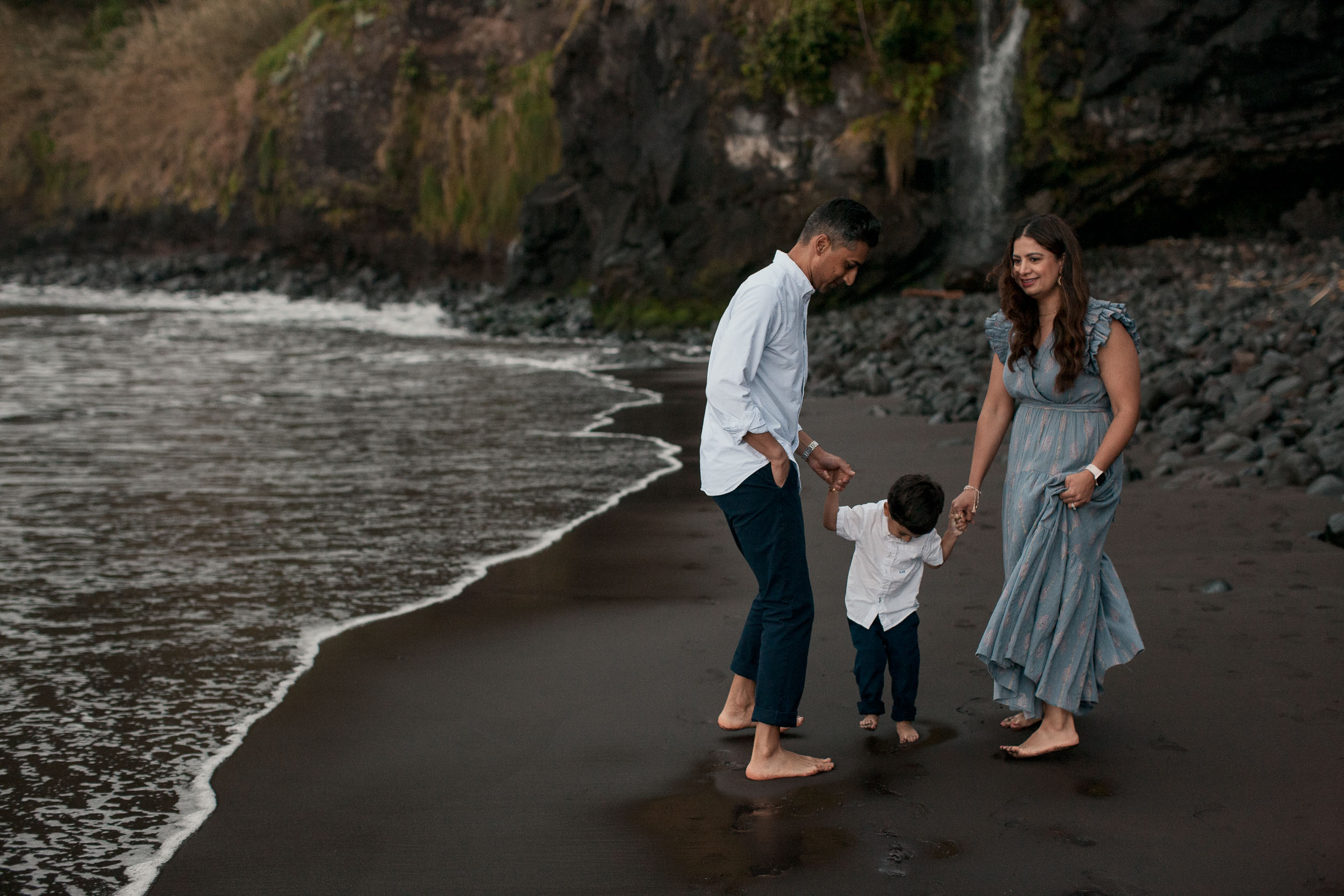Family walking along Seixal black sand shoreline, parents looking down at boy, small waterfall visible on cliff behind, Madeira