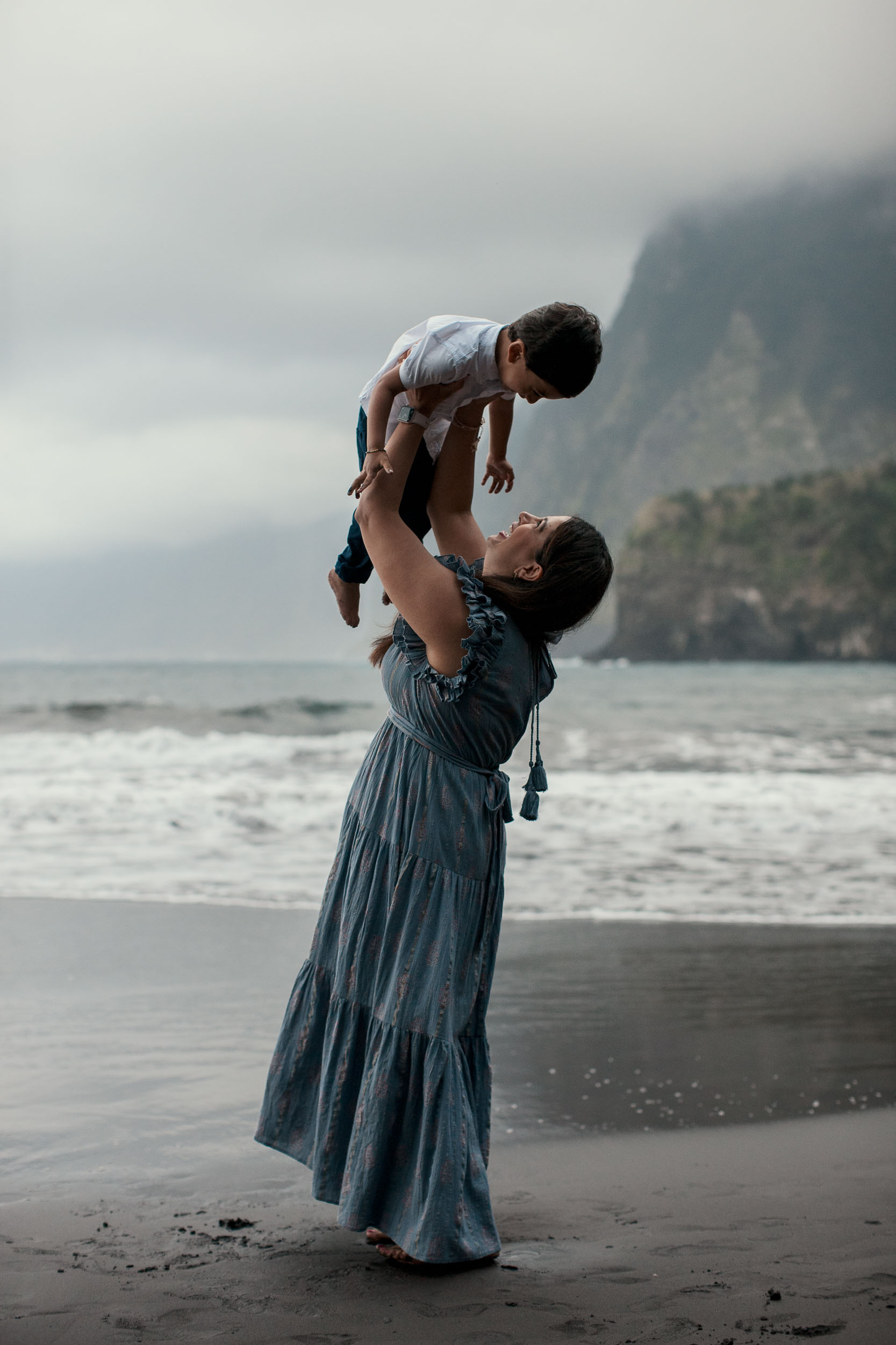 Mother lifting son high overhead on Seixal black sand beach, he looking down at her, dramatic misty cliffs and Atlantic behind, Madeira