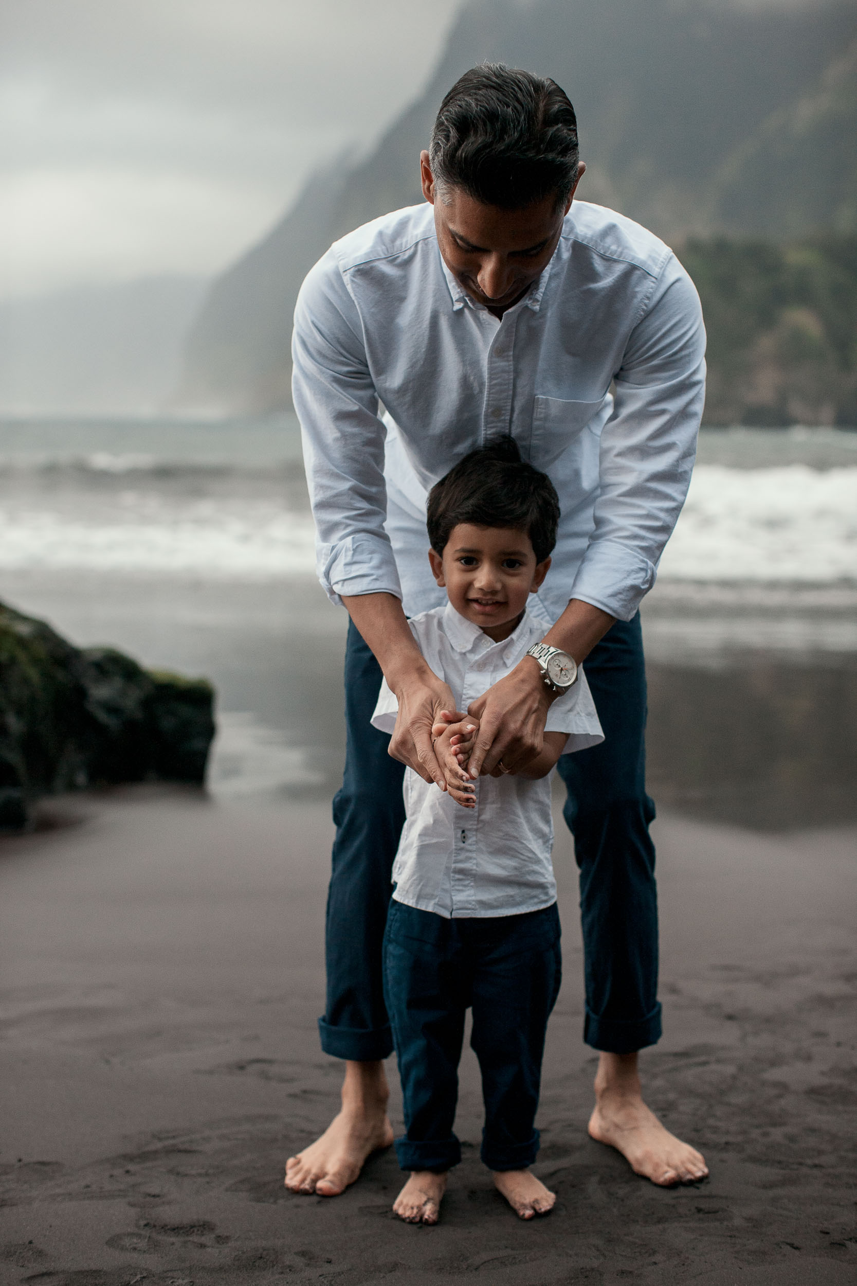 Father standing behind son on Seixal black sand beach, hands clasped over the boy's, both barefoot, dramatic cliffs and breaking waves behind, Madeira