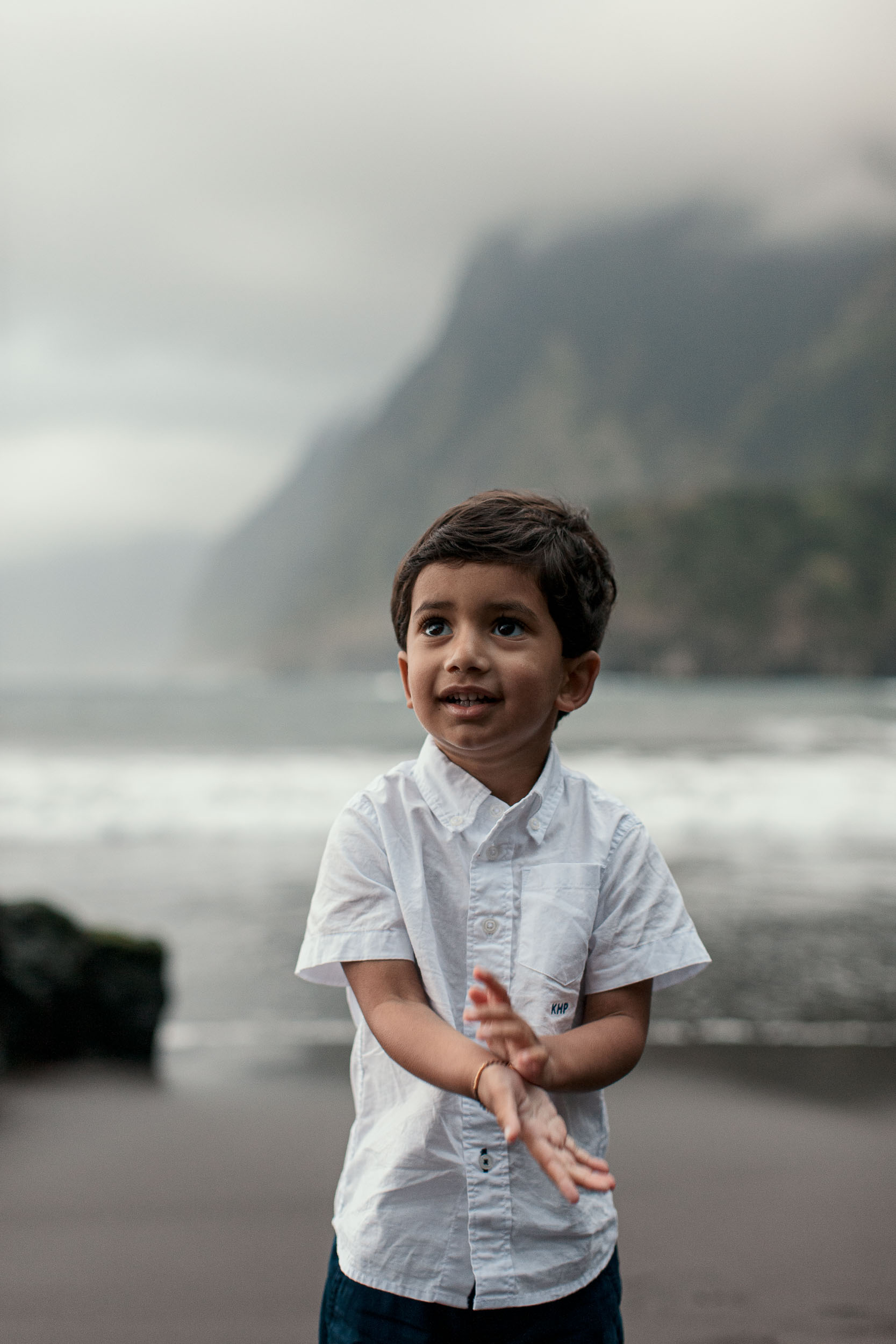 Boy standing alone on Seixal black sand beach looking to one side, dramatic misty cliffs and breaking waves behind, Madeira