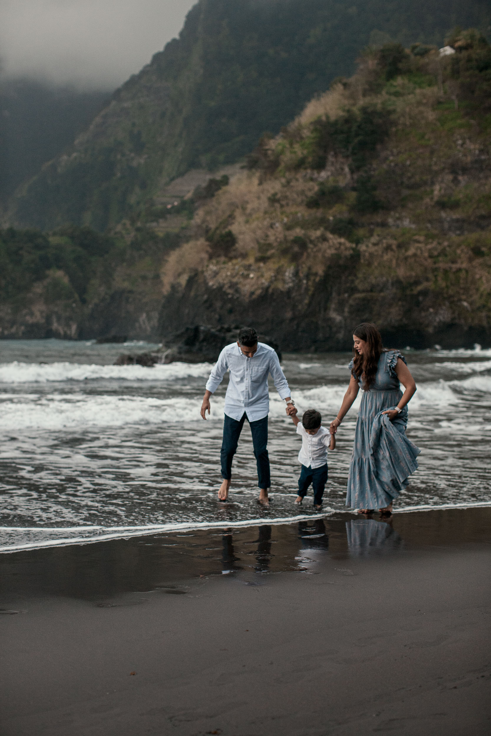 Family of three playing at the Seixal shoreline, father bending toward boy, mother watching and smiling, dramatic cliffs behind, Madeira