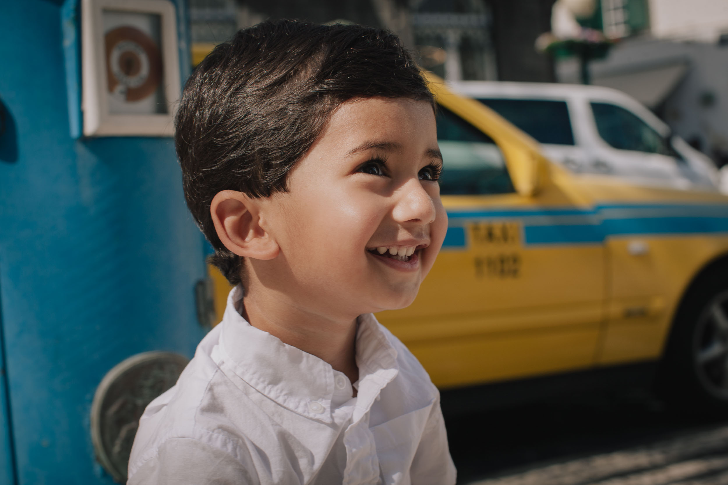 Close-up of boy laughing and looking sideways, blue Portuguese letter box beside him, yellow taxi behind, Funchal street, Madeira