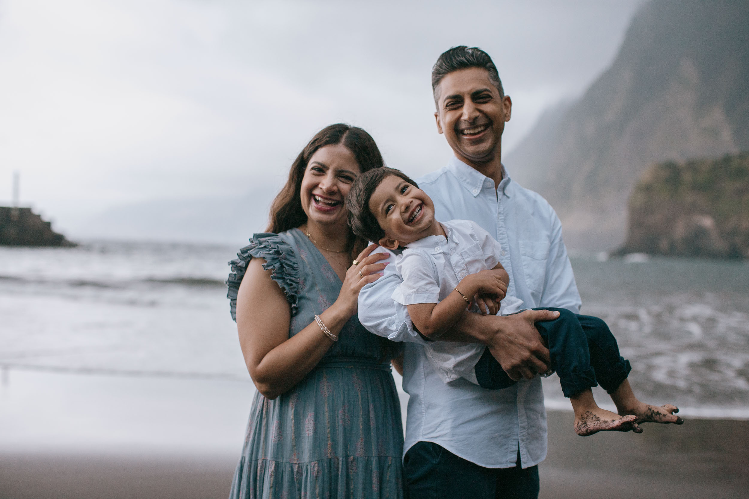 All three laughing together on Seixal black sand beach, father holding broadly laughing boy, mother beside them, Seixal cliffs behind, Madeira