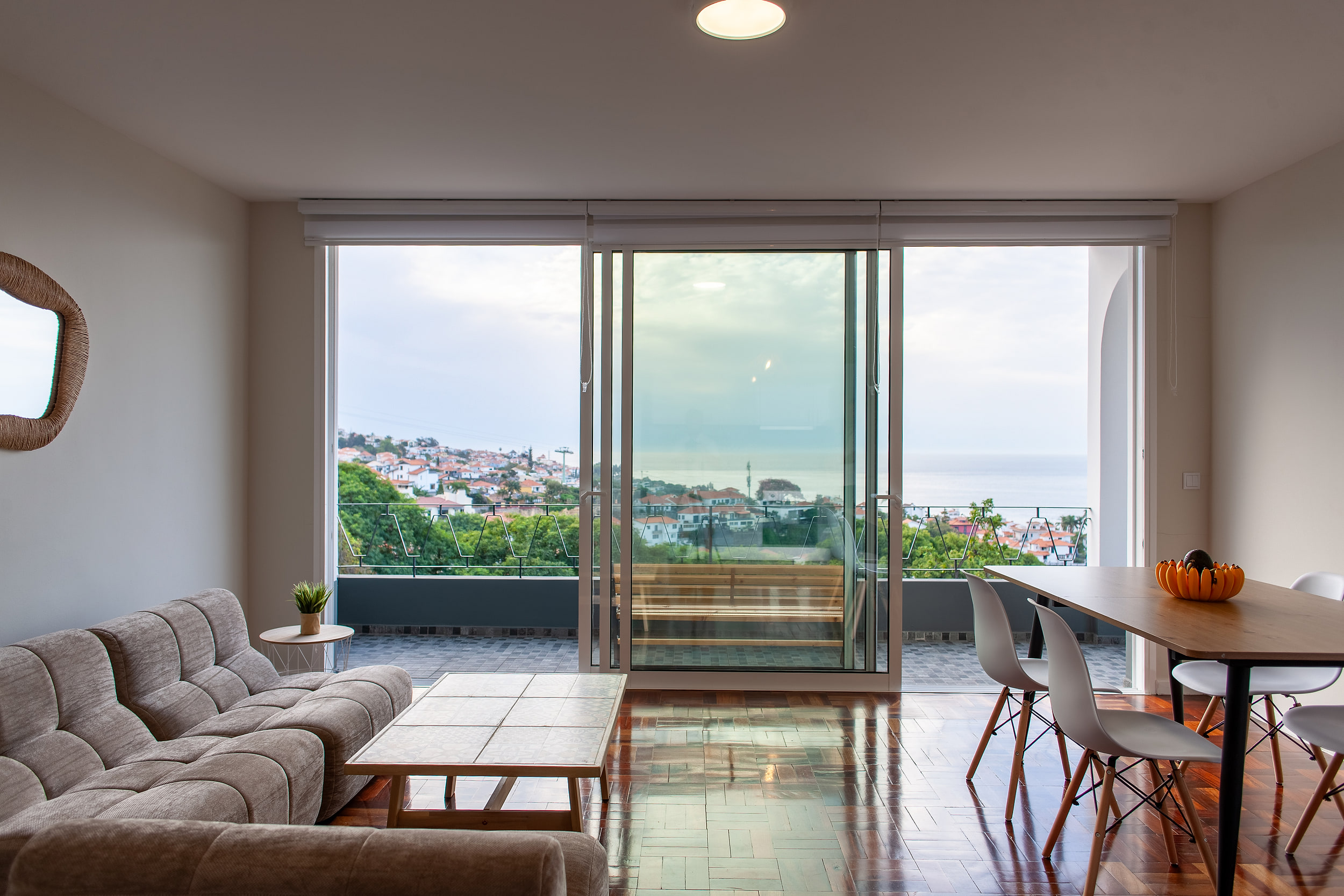 Living room facing the sliding glass doors, Funchal rooftops and the Atlantic outside, a wicker mirror on the wall
