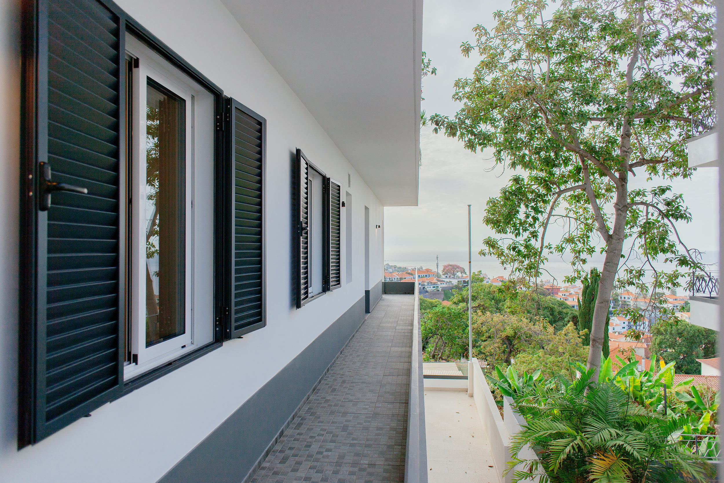 Outdoor walkway along the building with dark grey shutters, a tree and the sea at the end