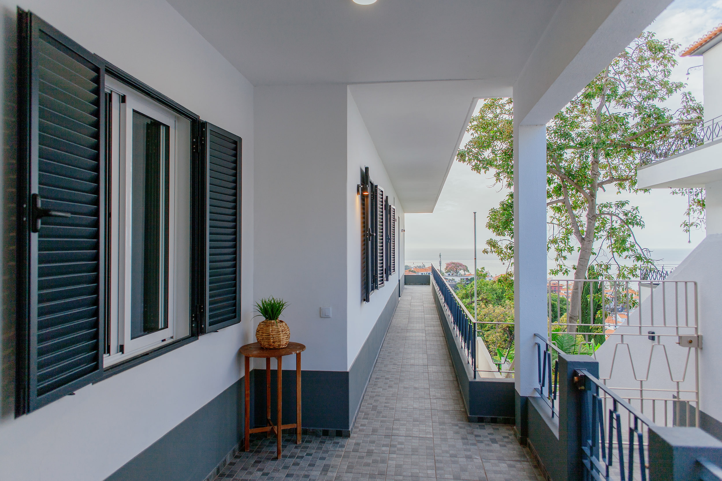 Covered walkway with grey shutters and a plant on a wooden stool, the sea at the far end