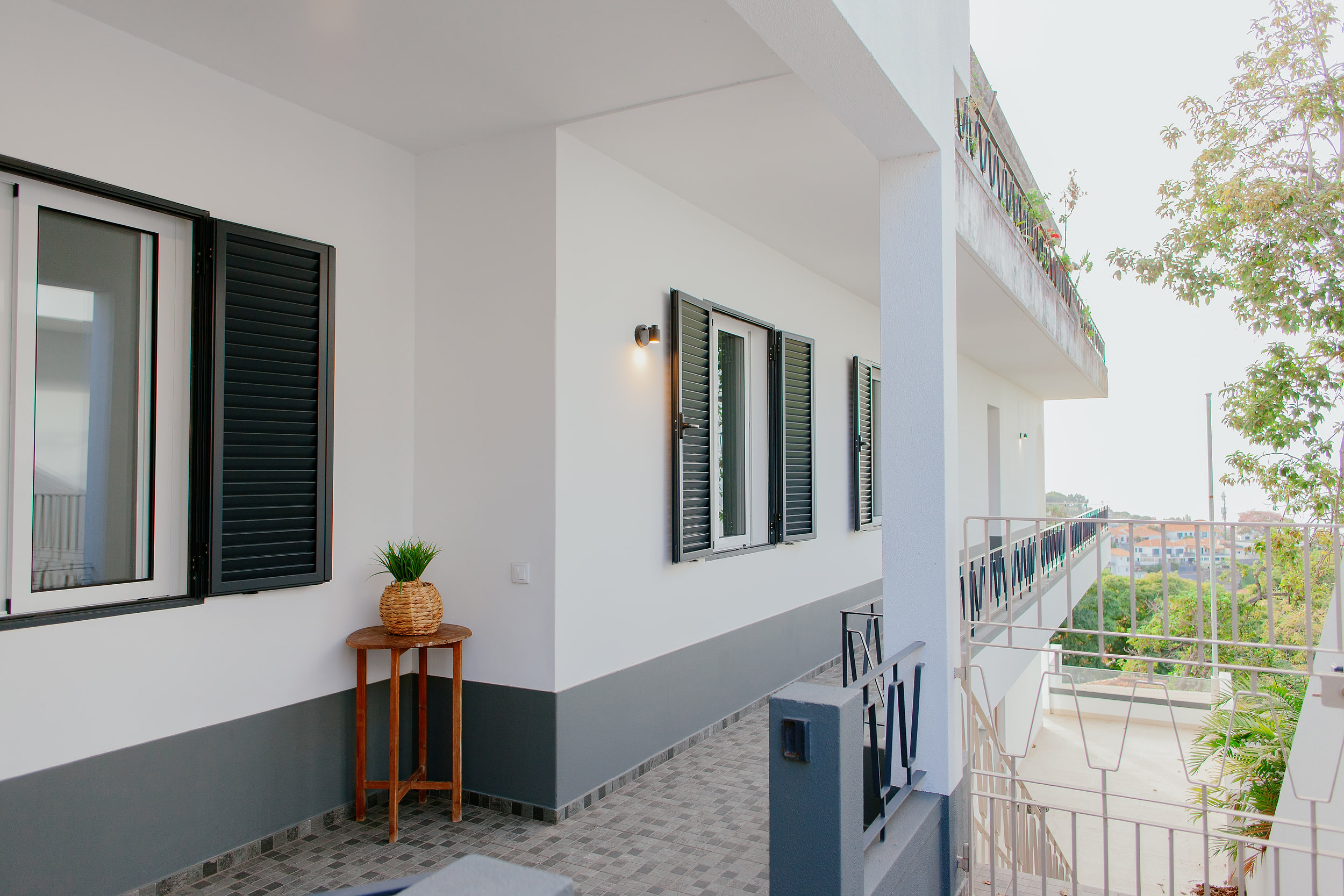 White facade with grey shutters and a terrace railing, trees and the ocean to the right