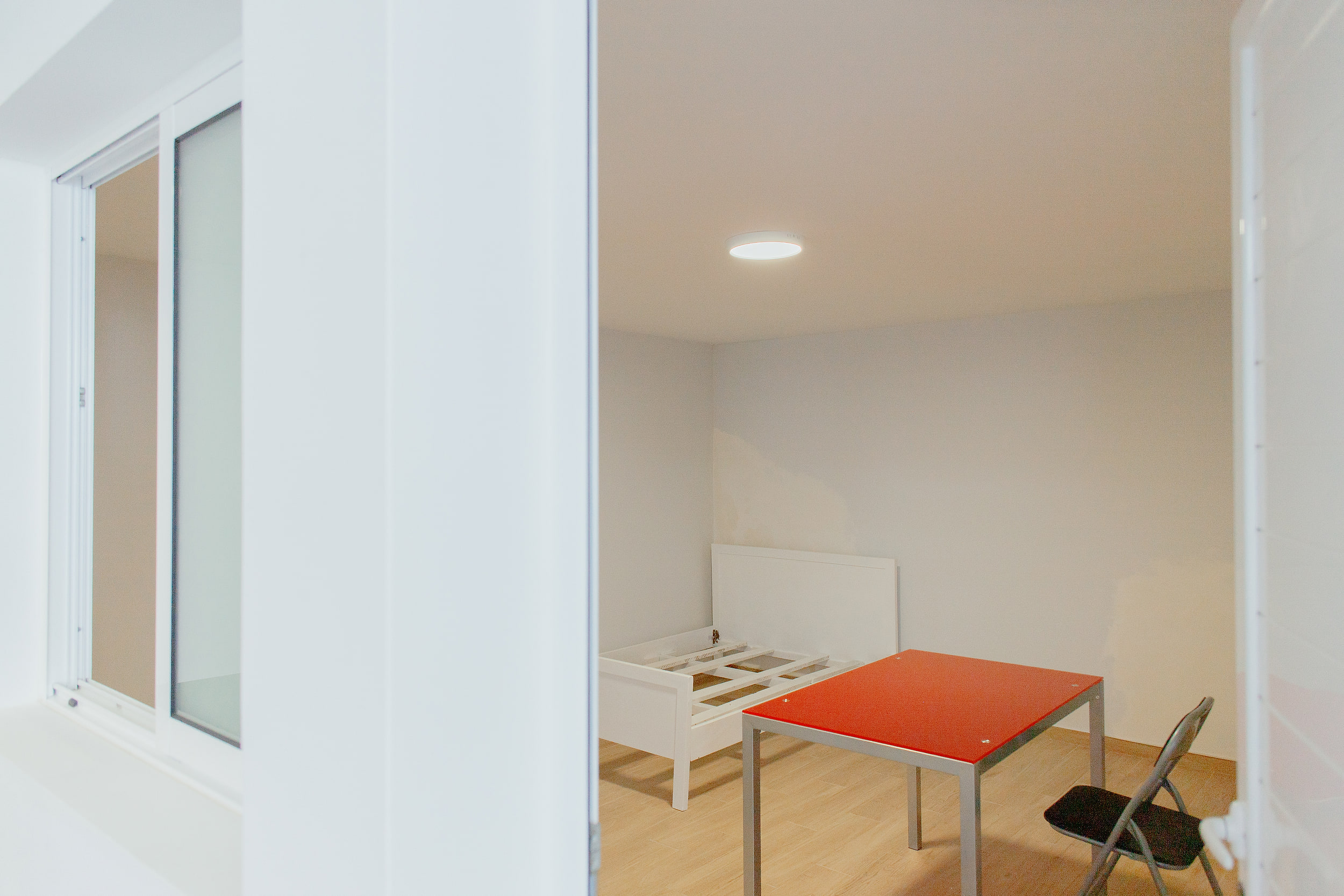 Lower room with a red table and a white bed frame, seen through the doorway