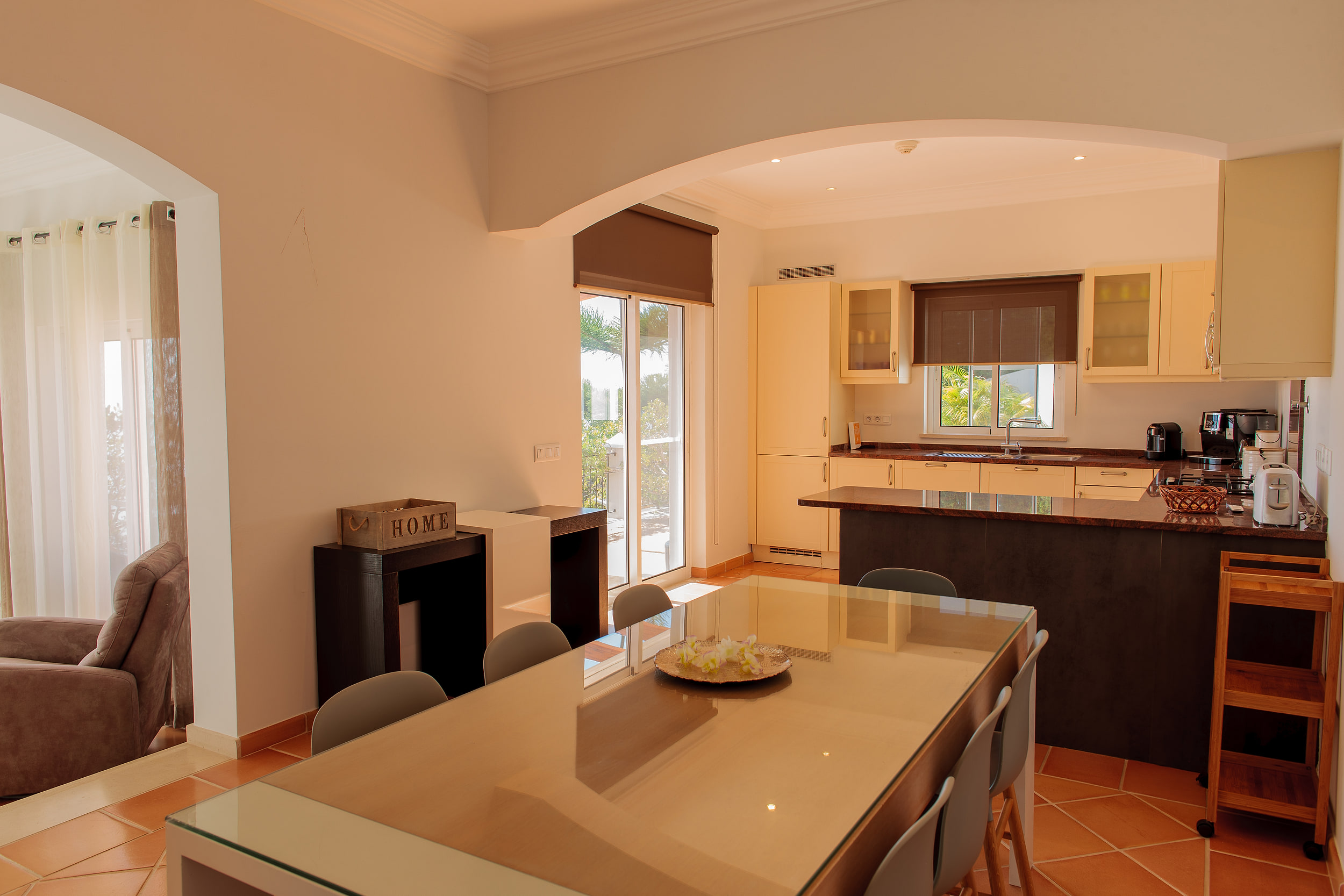 Open kitchen behind an arch, a dining table with grey chairs on terracotta floor tiles in sunlight