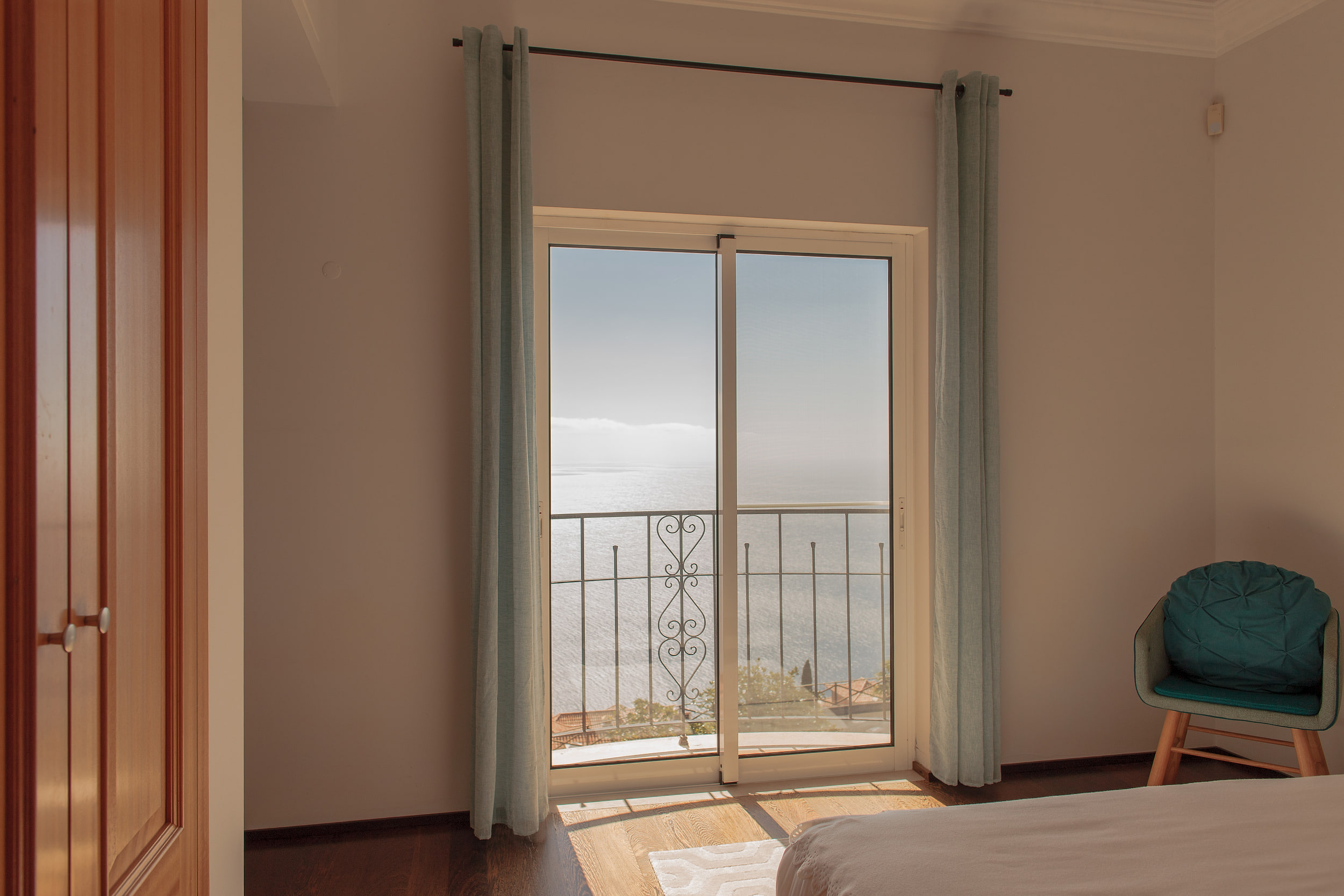 Bedroom with a sliding door onto a small balcony over the sea, teal curtains and an armchair