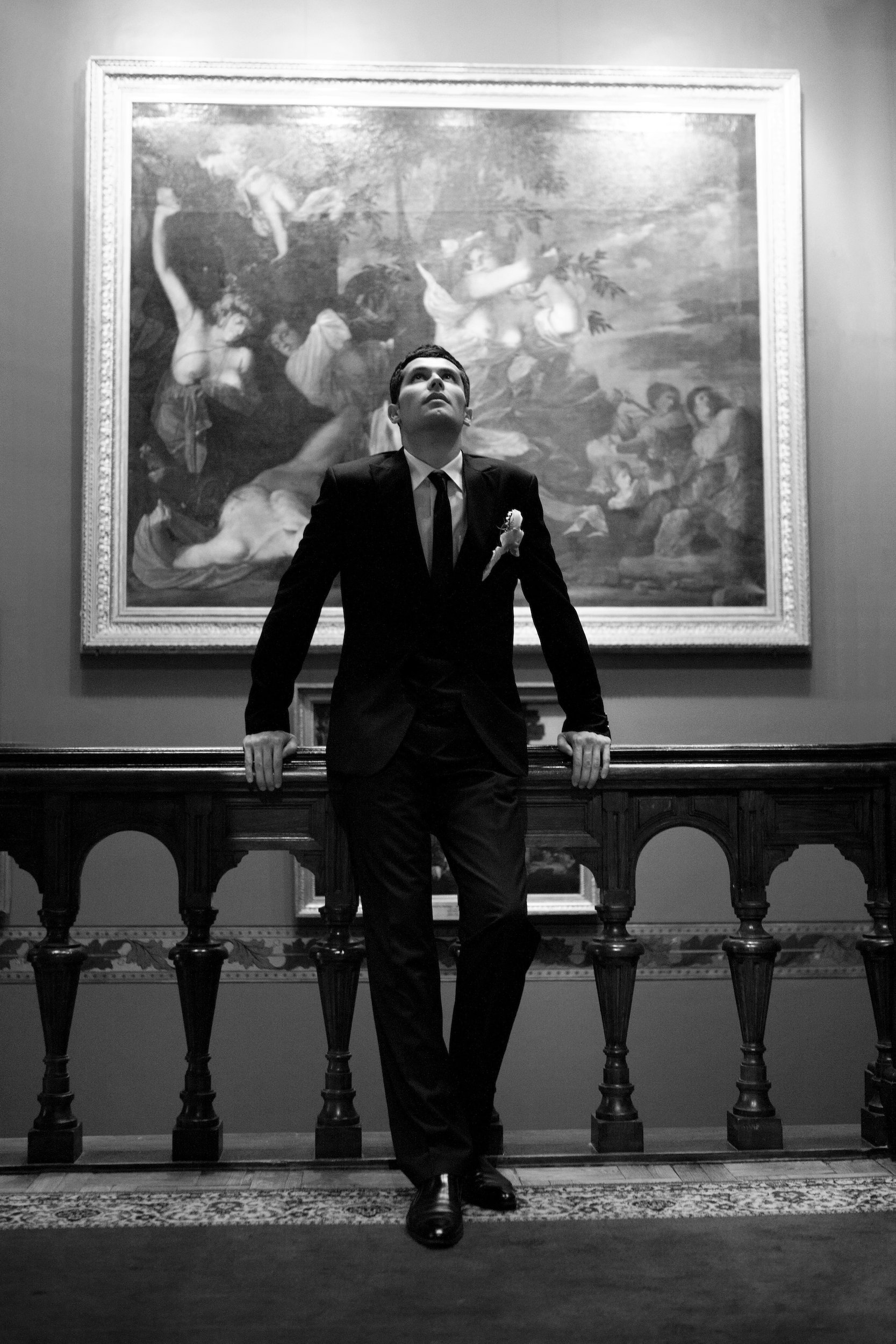 Groom in a dark suit leaning on a stone balustrade indoors, black and white